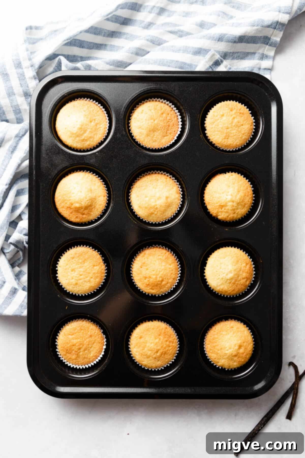 An overhead shot of a 12-hole cupcake baking tray, showcasing a dozen golden-brown vanilla cupcakes, fresh from the oven and perfectly baked.