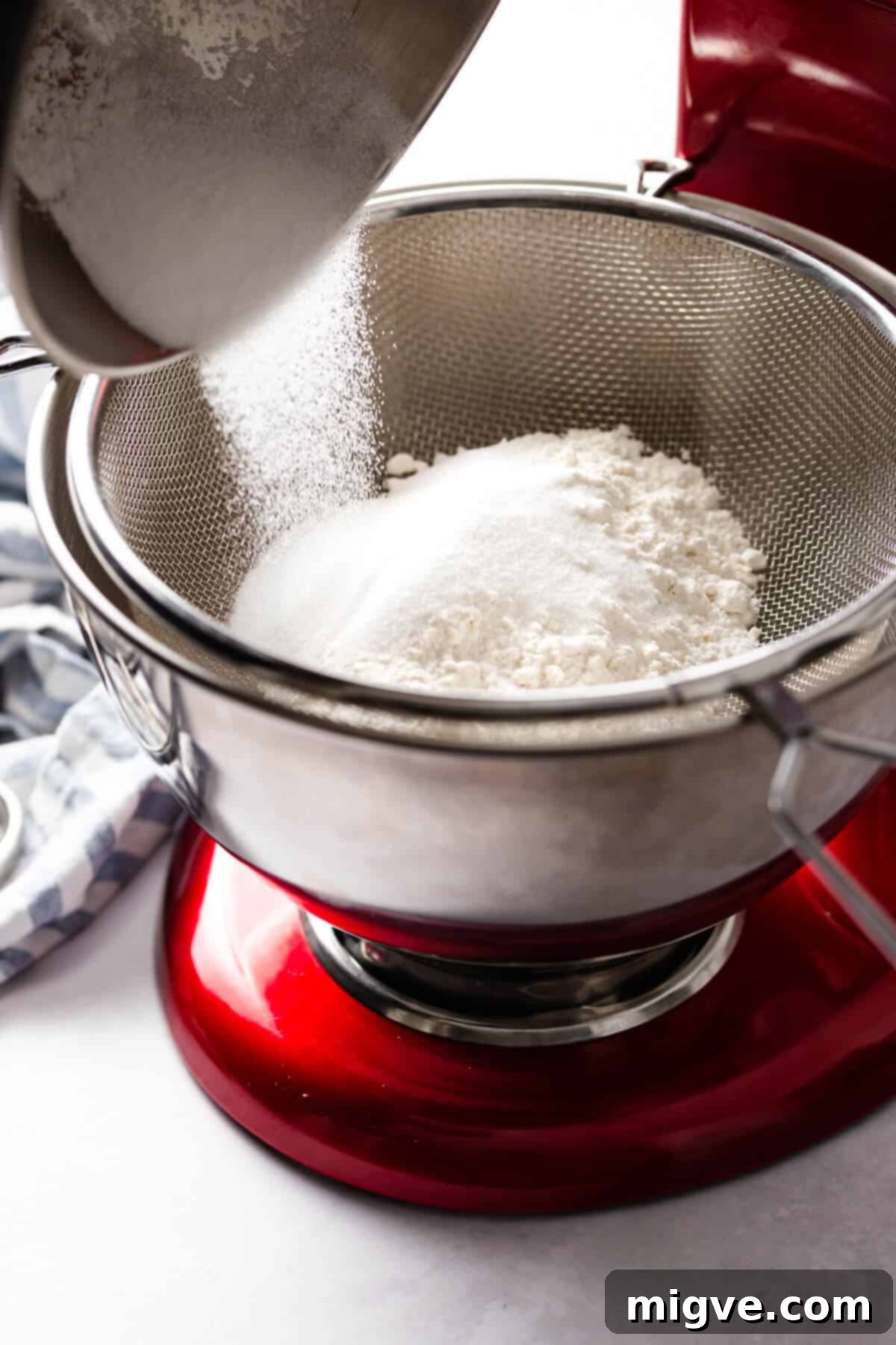 A side-angle photo capturing sugar being carefully added through a metal sieve into a bowl filled with flour, illustrating the crucial sifting step for smooth cupcake batter.