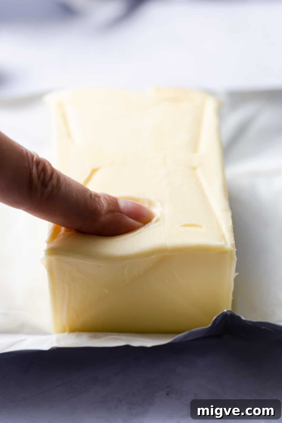 A side-angle shot of an index finger gently pressing into a block of butter, demonstrating the ideal room temperature consistency with a clear indent.