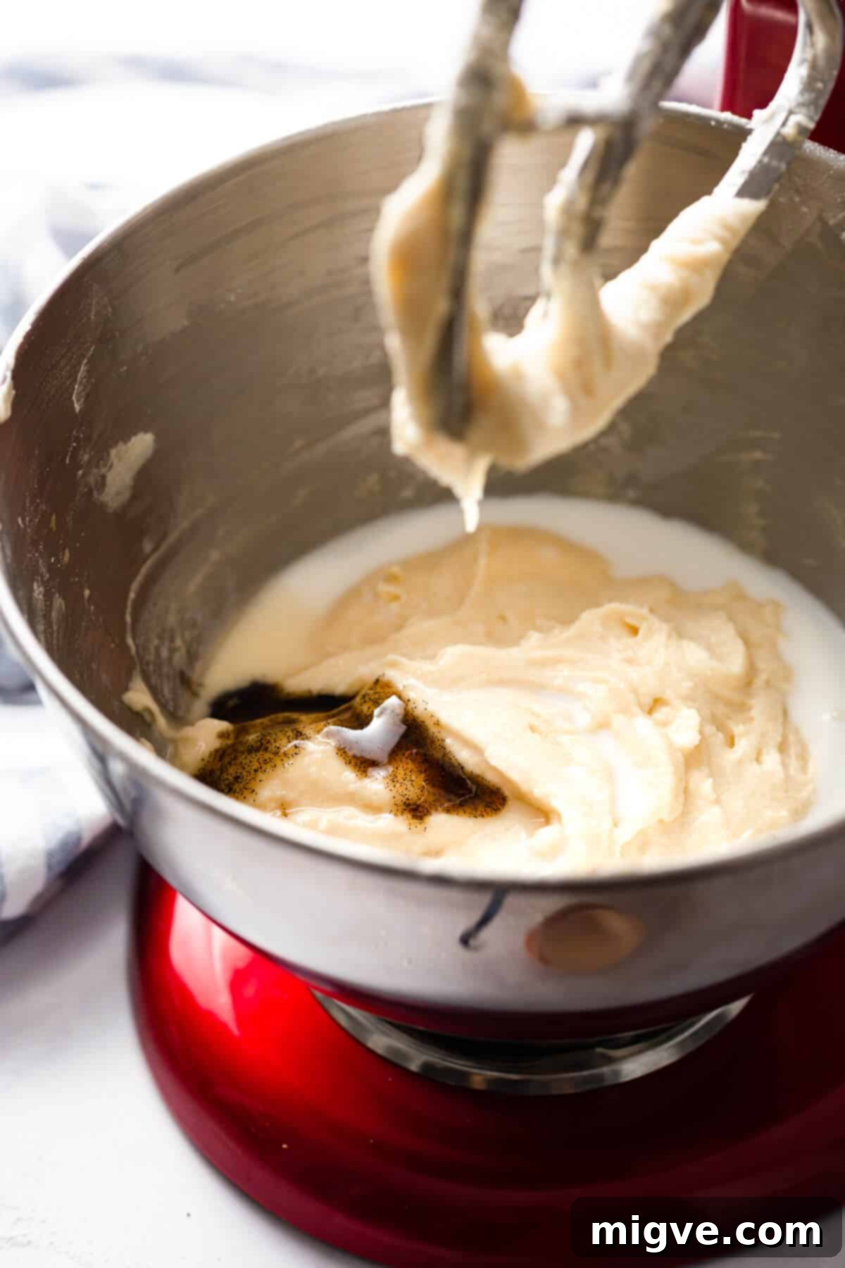 A side-angle shot showing a stream of vanilla extract and milk being carefully poured into a mixing bowl containing fluffy cupcake batter.
