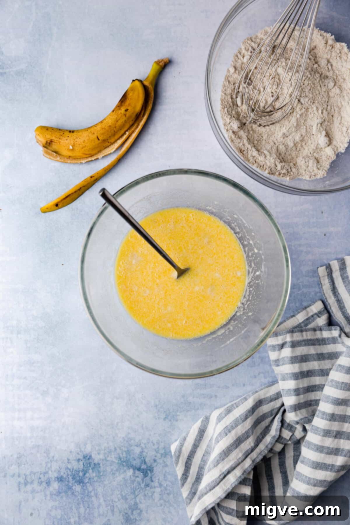 Overhead image showing clear glass bowls filled with wet and dry ingredients, alongside a banana peel, signifying preparation