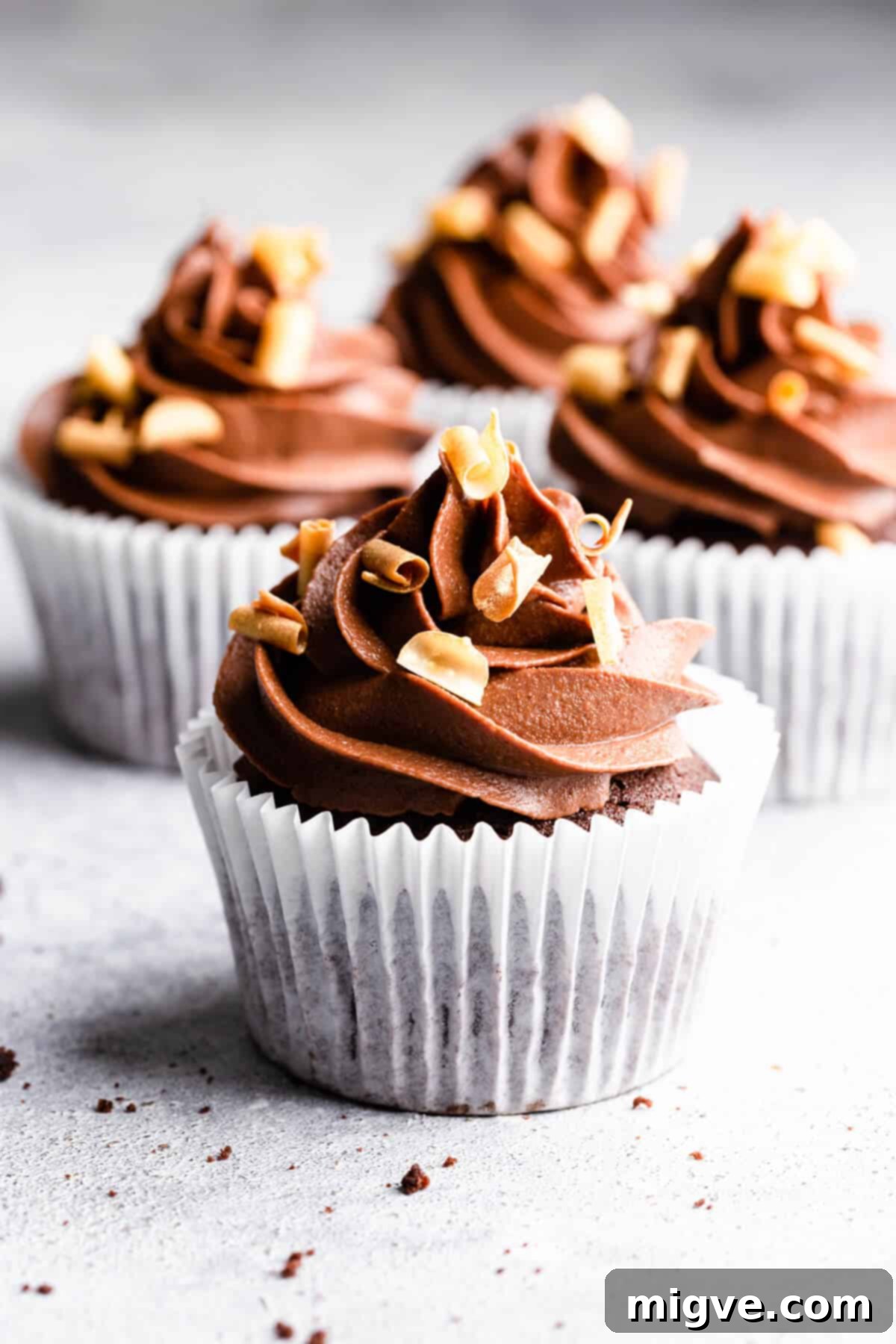 Straight ahead angle close up of four chocolate cupcakes with chocolate buttercream and delicate chocolate curls on top, ready to be enjoyed.