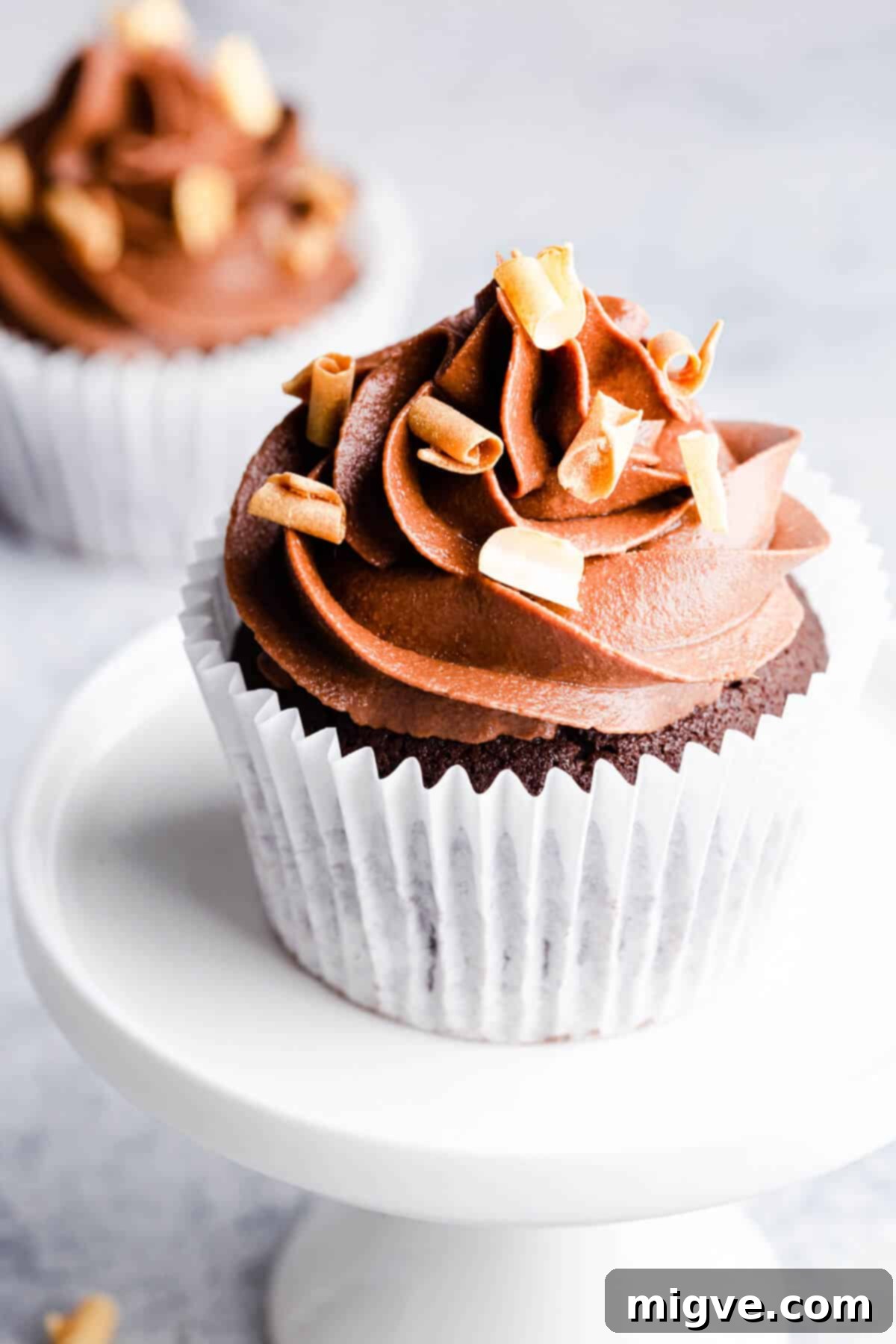 A side angle photo of a single chocolate cupcake on a small white cupcake stand, showcasing its perfect frosting swirl.