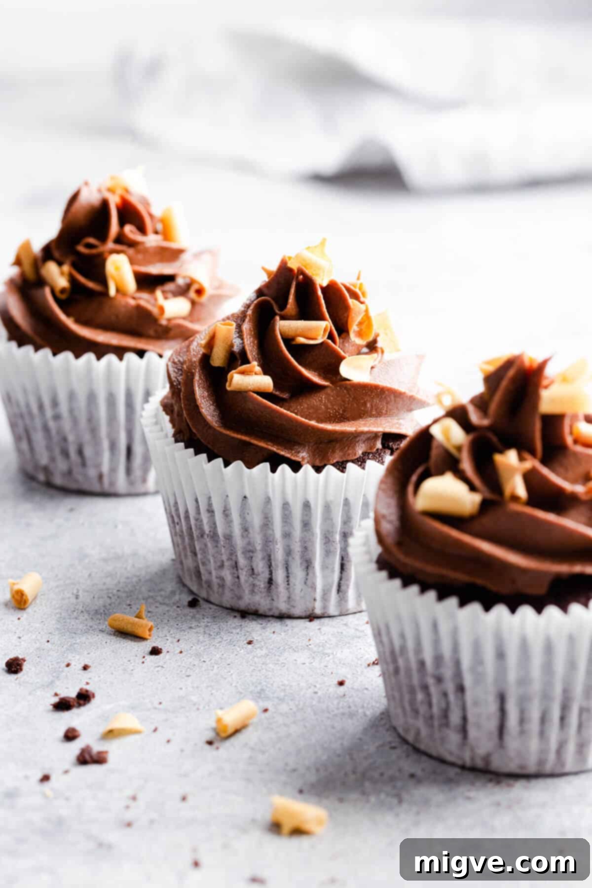 Side angle photo of 3 chocolate cupcakes, with one in sharp focus, showcasing its delightful frosting and crumb.