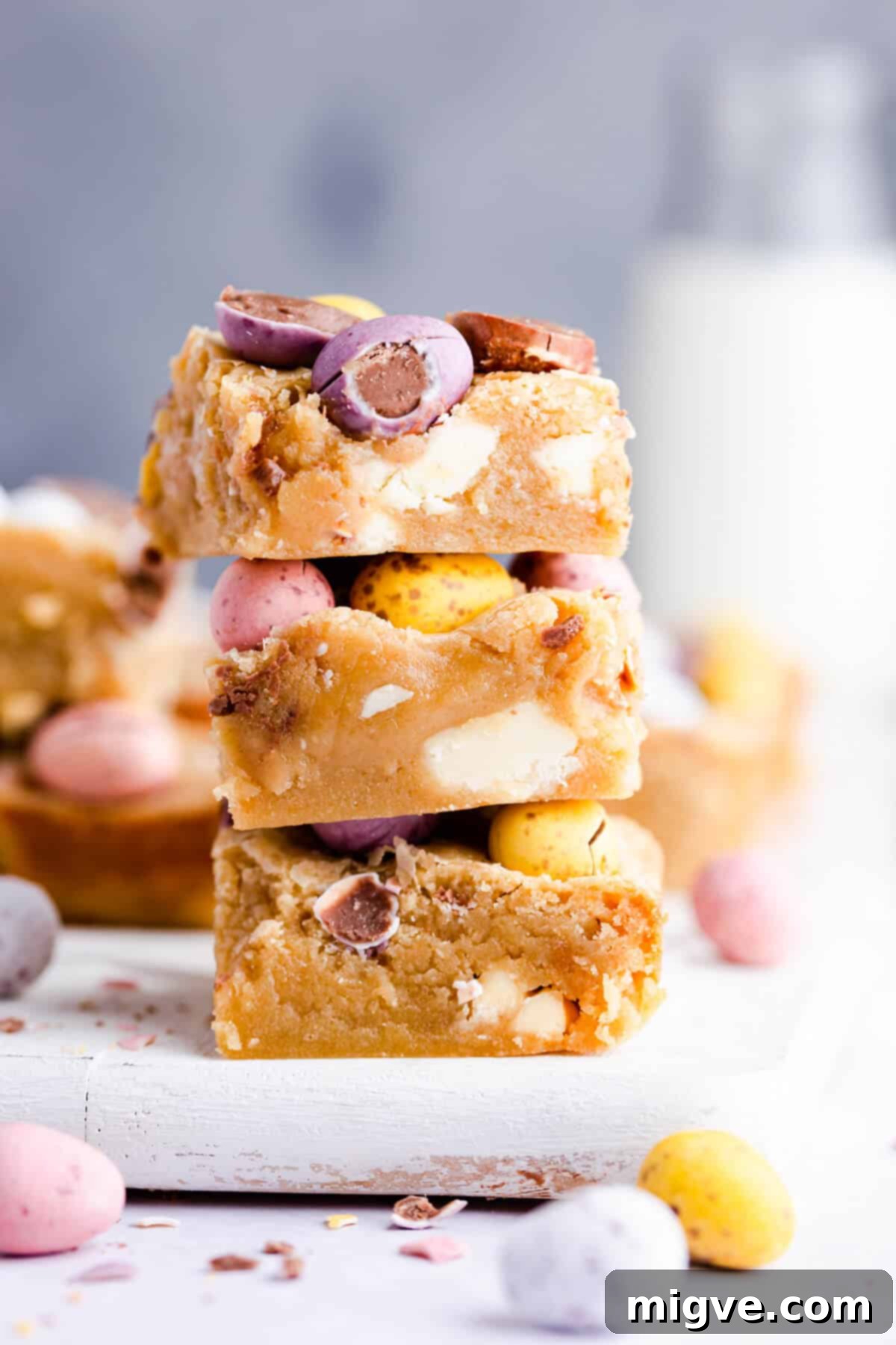 Speckled Spring Blondies 2 A stack of three golden mini egg blondies with white chocolate chunks and colorful mini eggs on top, viewed from a side angle.