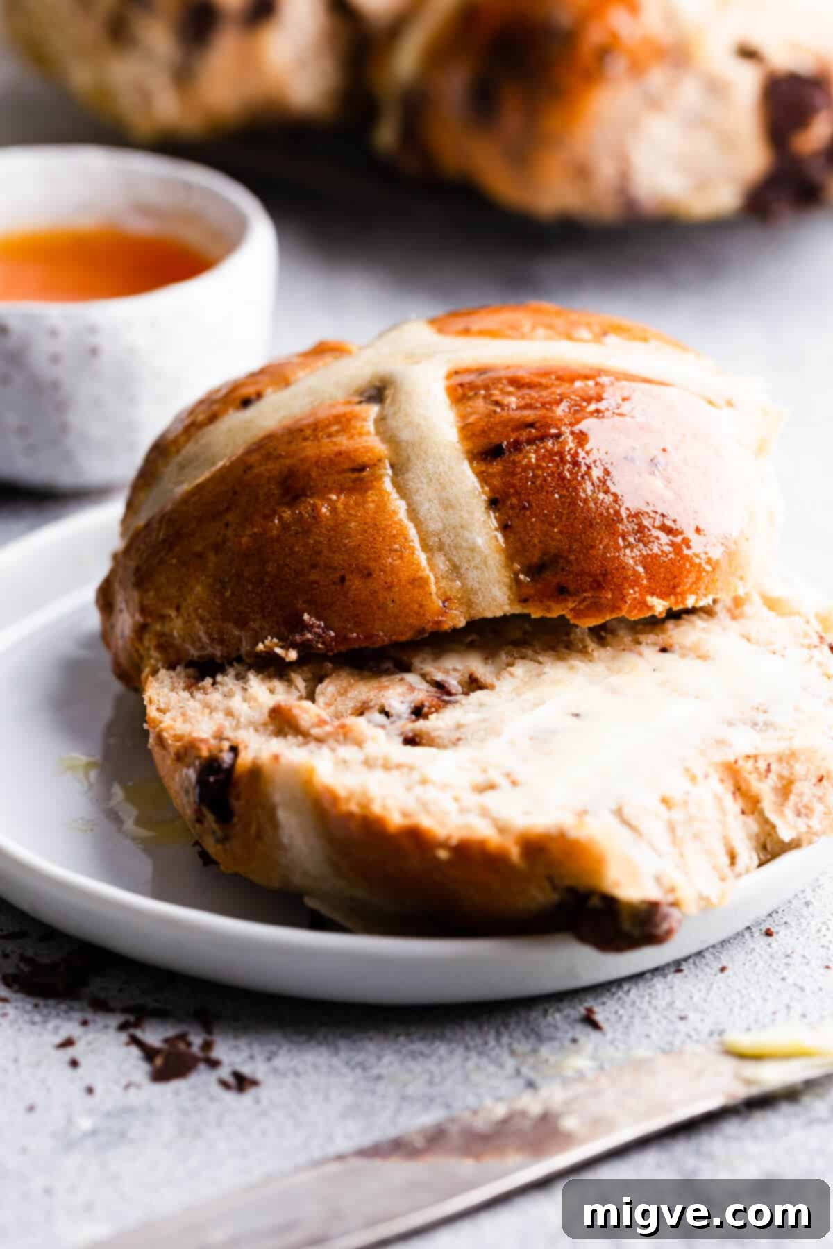 A close-up side view of a chocolate chip hot cross bun, perfectly glazed and sliced, with butter melting into its fluffy interior.