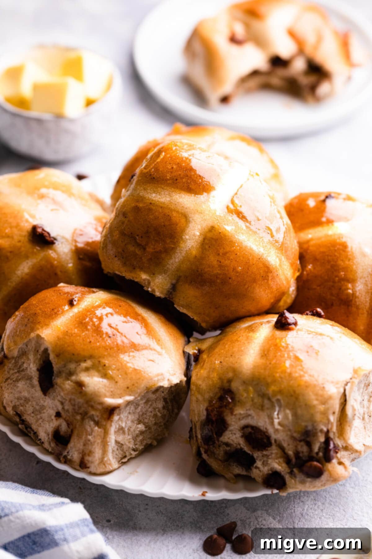A person is shown brushing a shiny apricot glaze onto warm, freshly baked hot cross buns with a pastry brush.