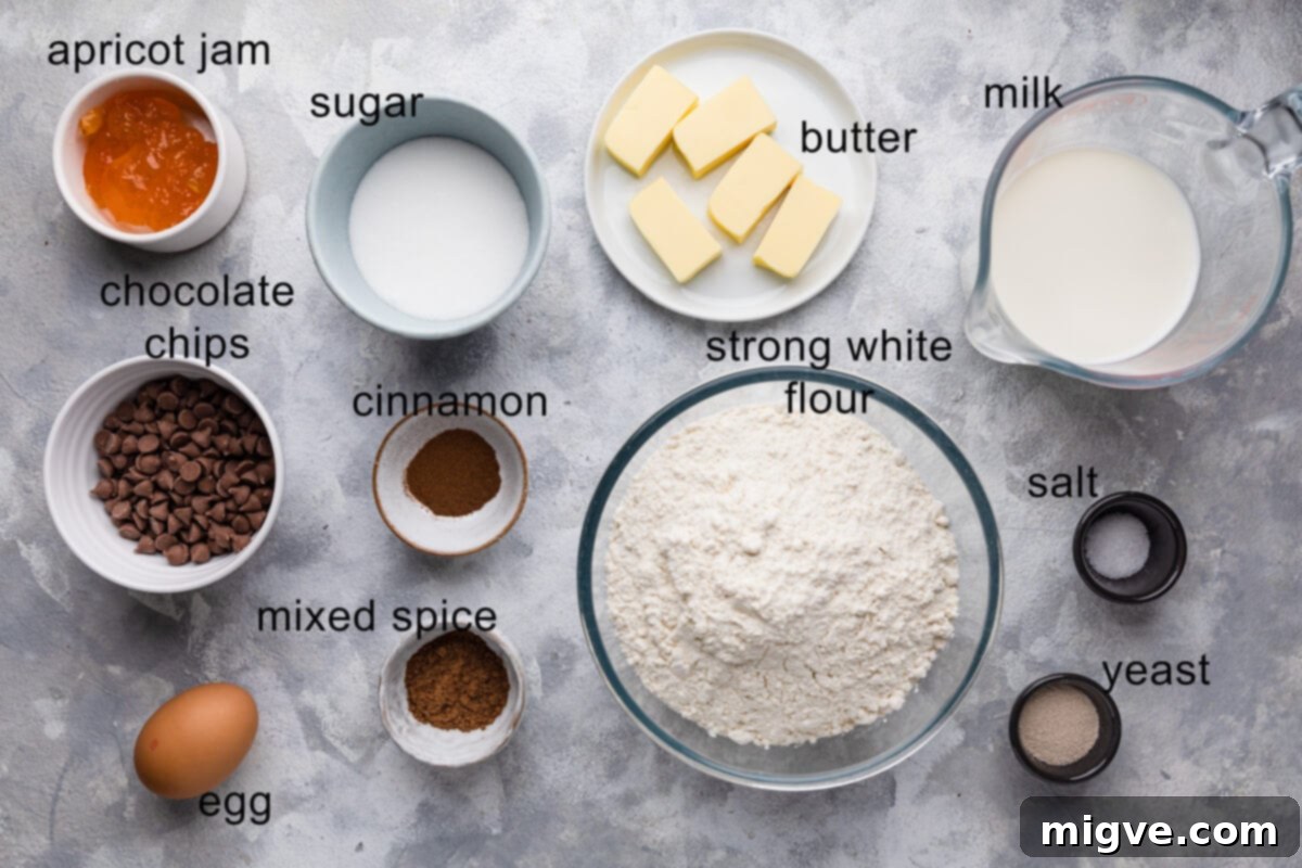 A top-down view of all the measured ingredients for chocolate chip hot cross buns laid out on a wooden surface with text labels, including flour, sugar, yeast, spices, butter, milk, egg, and chocolate chips.