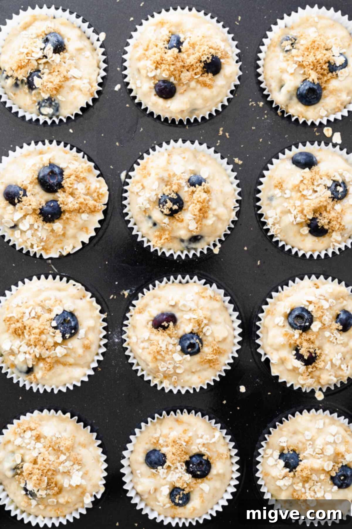 Overhead view of a baking tin, each cavity filled to the top with banana and blueberry muffin batter in paper cases, sprinkled with topping.