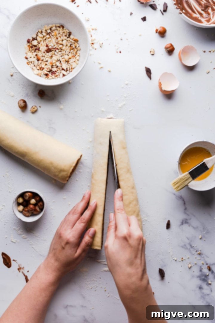 Top-down view of a person slicing a chilled, rolled babka dough log lengthways into two long, filled strands.