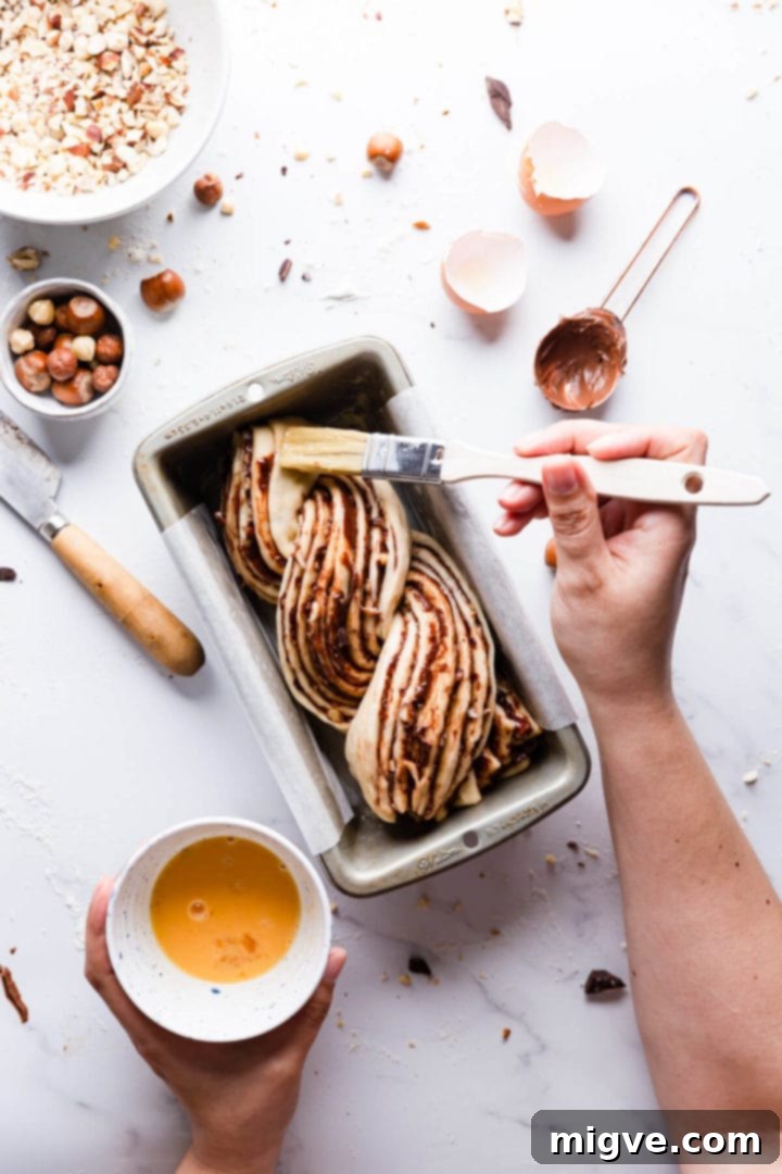 Overhead view of a chocolate hazelnut babka in a loaf pan being carefully brushed with an egg wash before baking.