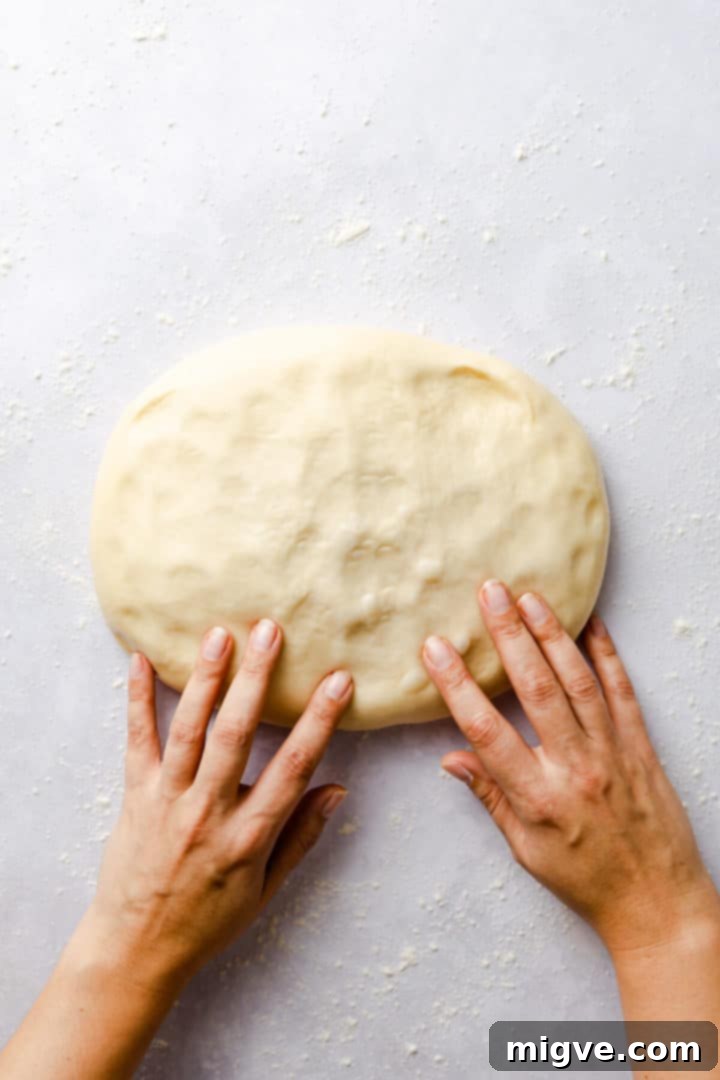 Top-down view of a person gently knocking the air out of soft, risen dough on a floured surface.