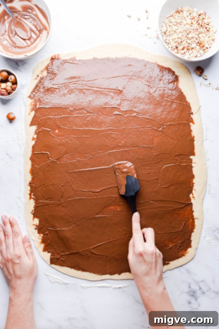 Top-down view of a person evenly spreading Nutella over the rectangular babka dough using a spatula.