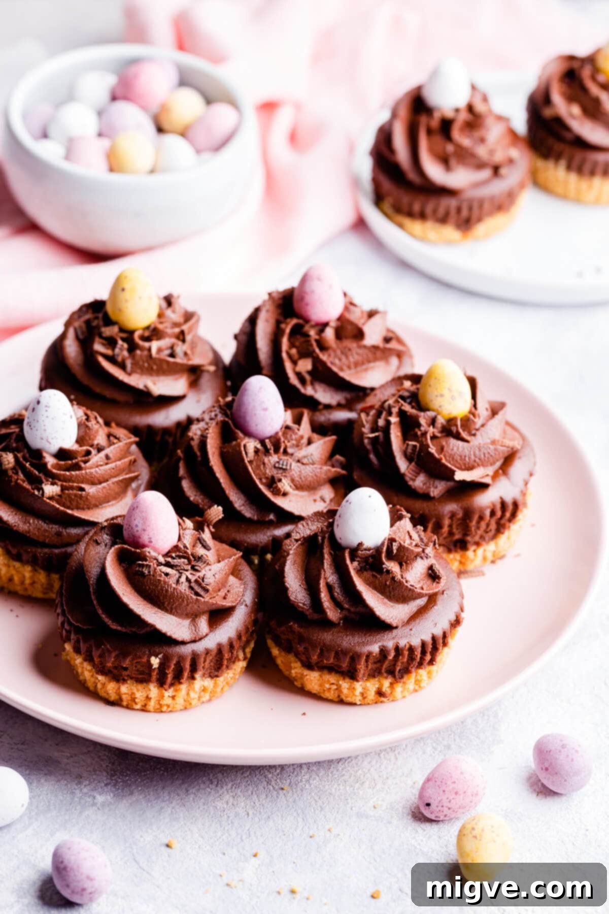 Four chocolate mini cheesecakes adorned with mini Easter eggs on a white plate, viewed from a 45-degree angle.