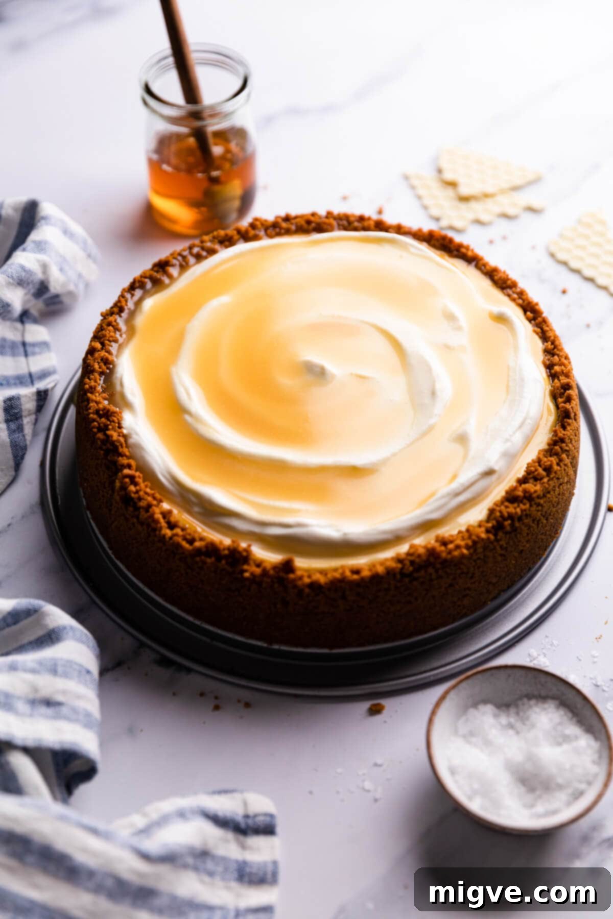 Velvet Honey Cheesecake 2 45 degree angle view at honey cheesecake topped with cream and honey, showcasing its rich, golden-brown salted honey drizzle and white sour cream against a crunchy ginger nut crust