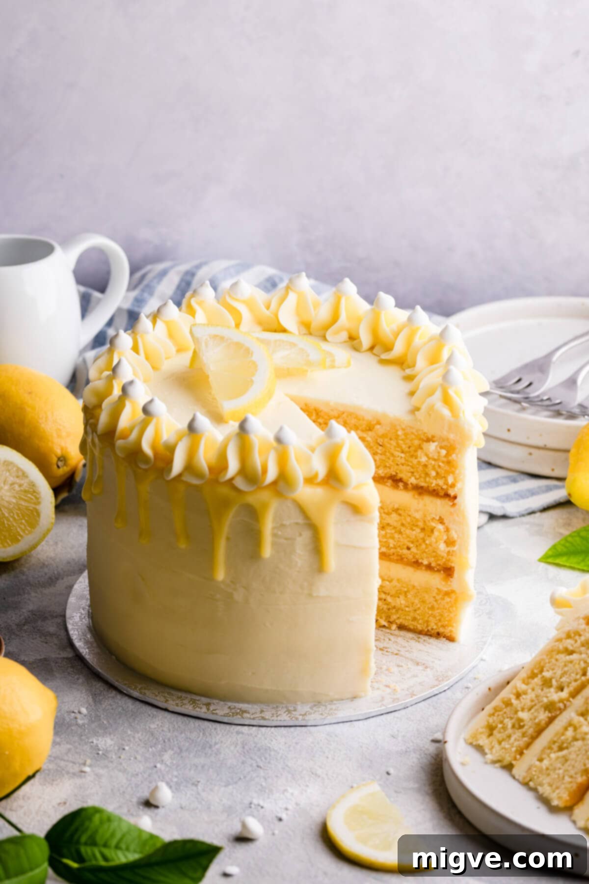 A stunning three-layer lemon and elderflower cake on a cake board with a perfect slice removed, revealing the delightful layers of sponge, buttercream, and lemon curd, adorned with a white chocolate ganache drip and fresh elderflowers.