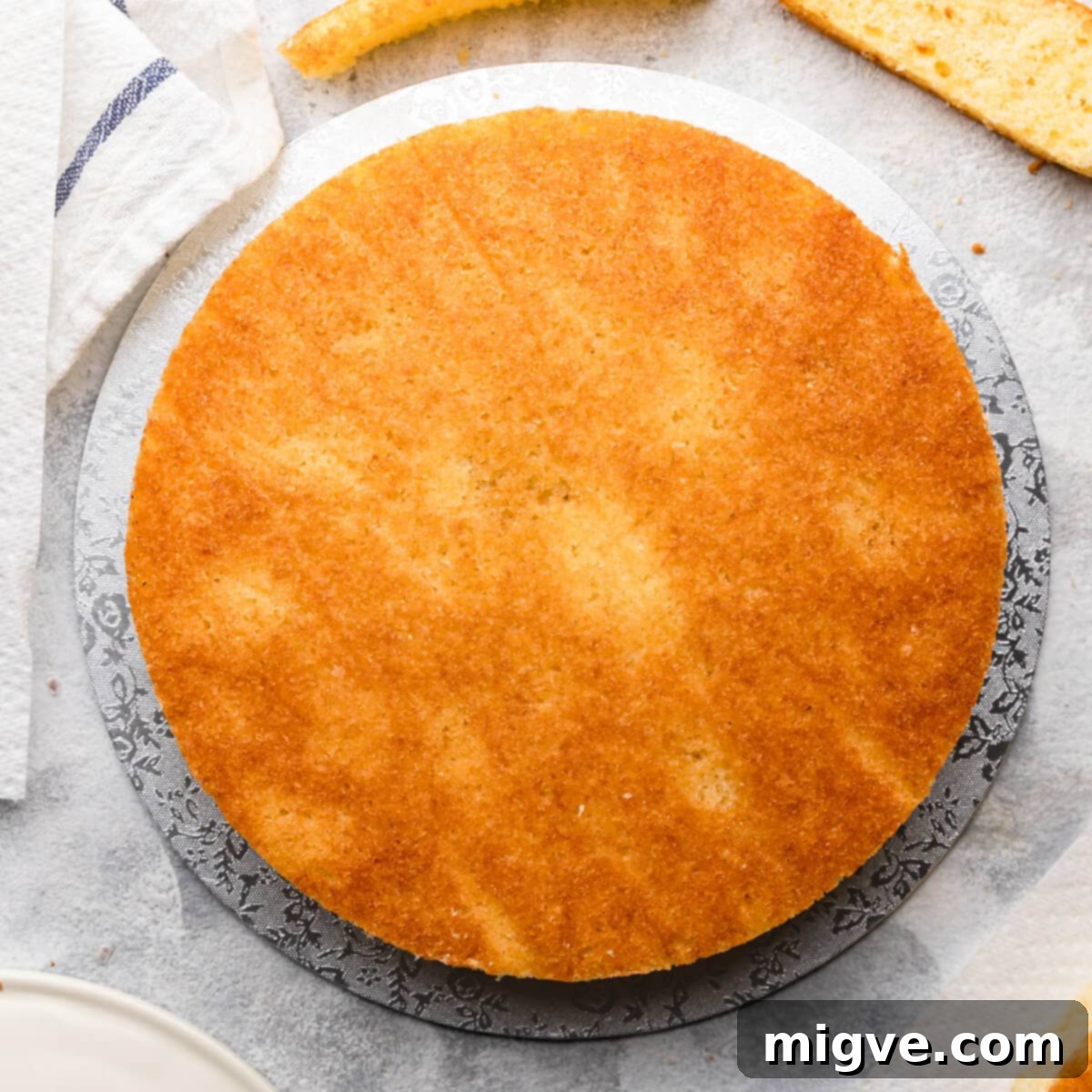 Top-down view of a cooled sponge cake with its edges neatly trimmed, placed on a cake board and brushed with a shimmering syrup, ready for the next layer.