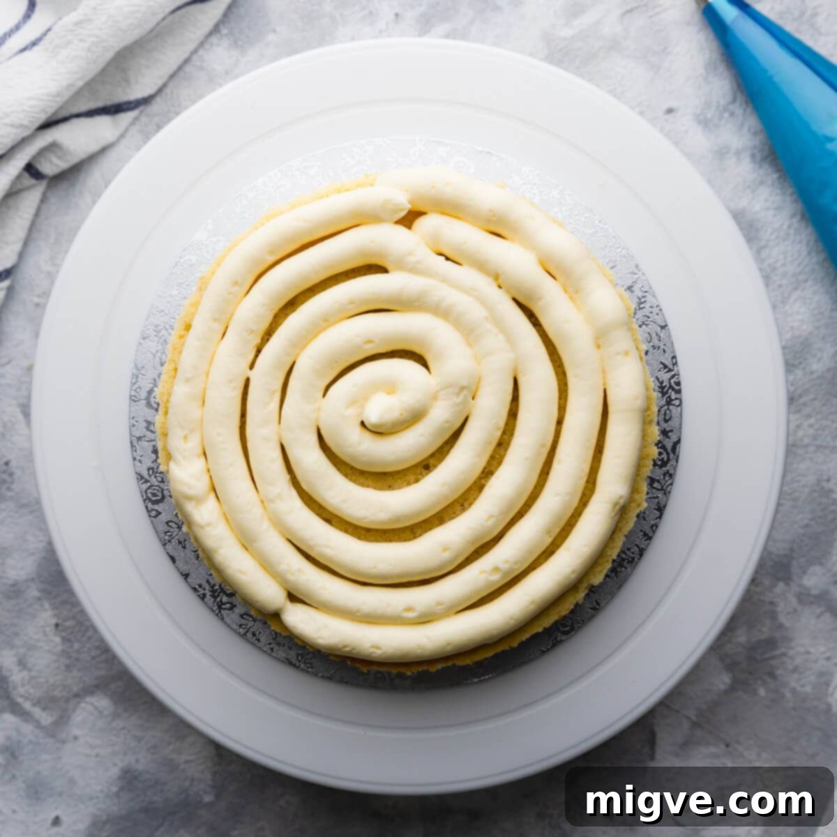 Overhead view of the first sponge cake layer topped with a generous, evenly piped layer of fluffy elderflower buttercream, creating a perfect base for the lemon curd.