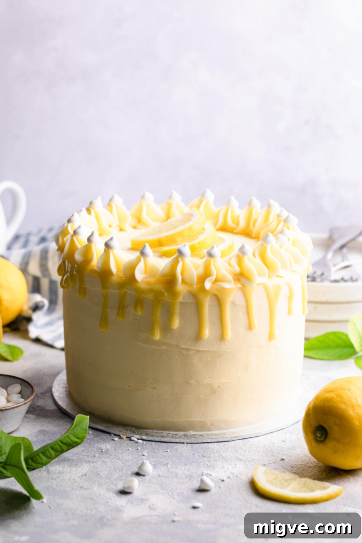 Straight-ahead view of the magnificent three-layer elderflower and lemon cake, showcasing its perfect white chocolate drip, delicate buttercream swirls, and fresh garnishes, ready to be served.