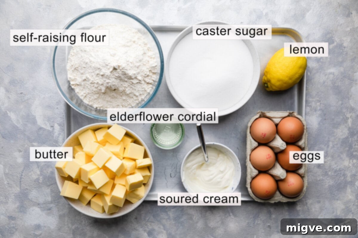 Overhead view of carefully arranged ingredients for the lemon and elderflower cake sponge, including butter, sugar, eggs, soured cream, elderflower cordial, flour, and a lemon, each labeled for clarity.