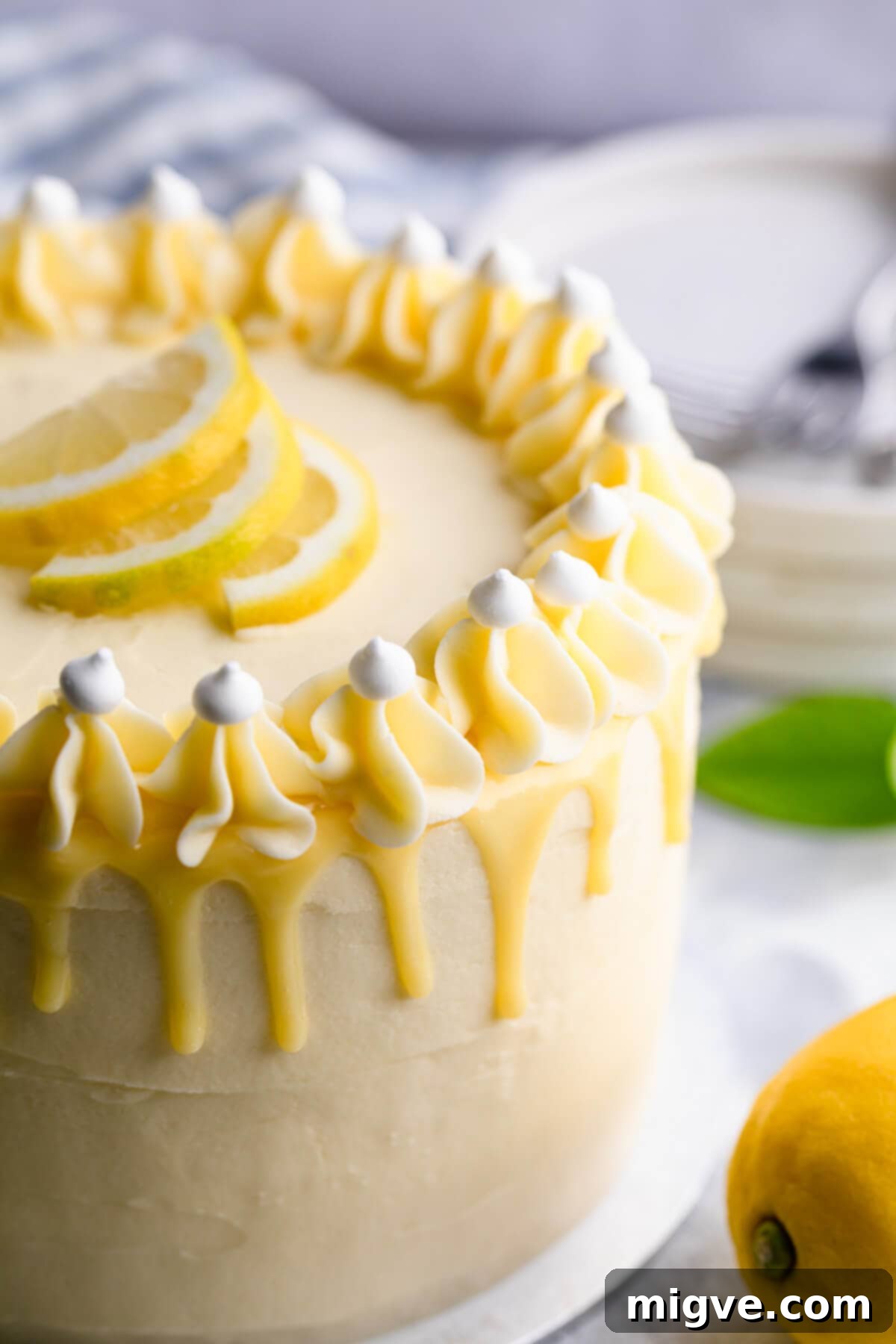 A 45-degree super close-up shot of the top of the lemon and elderflower cake, highlighting the elegant white chocolate ganache drip cascading down the side and the intricate piped buttercream decoration.