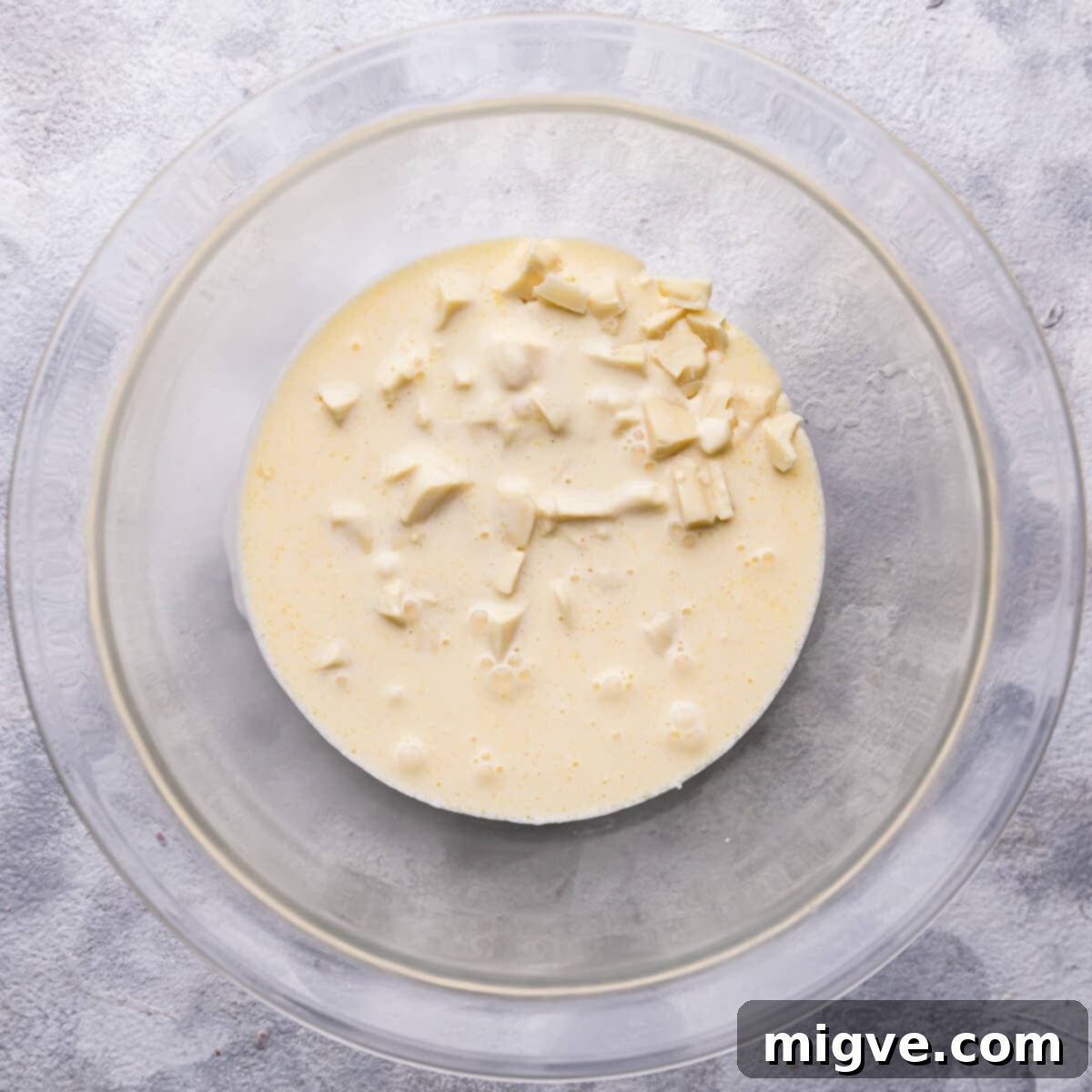 Overhead view of hot double cream and melted butter being slowly poured over the finely chopped white chocolate in a heatproof bowl, creating the initial stage of ganache.