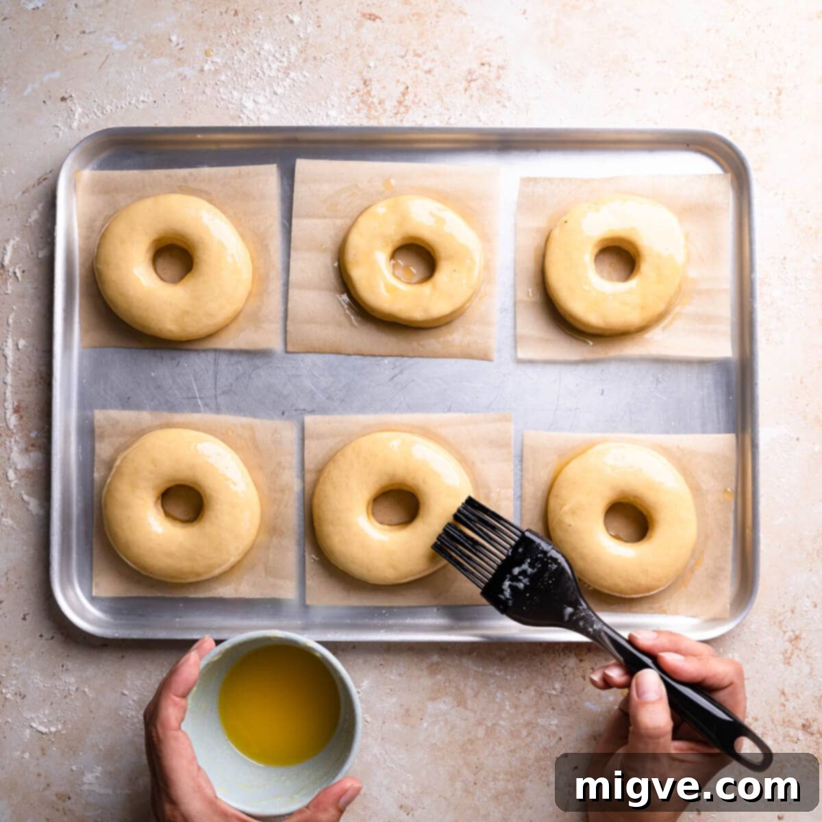 Unbaked doughnuts neatly arranged on a baking sheet, brushed with melted butter, and ready for their second rise.