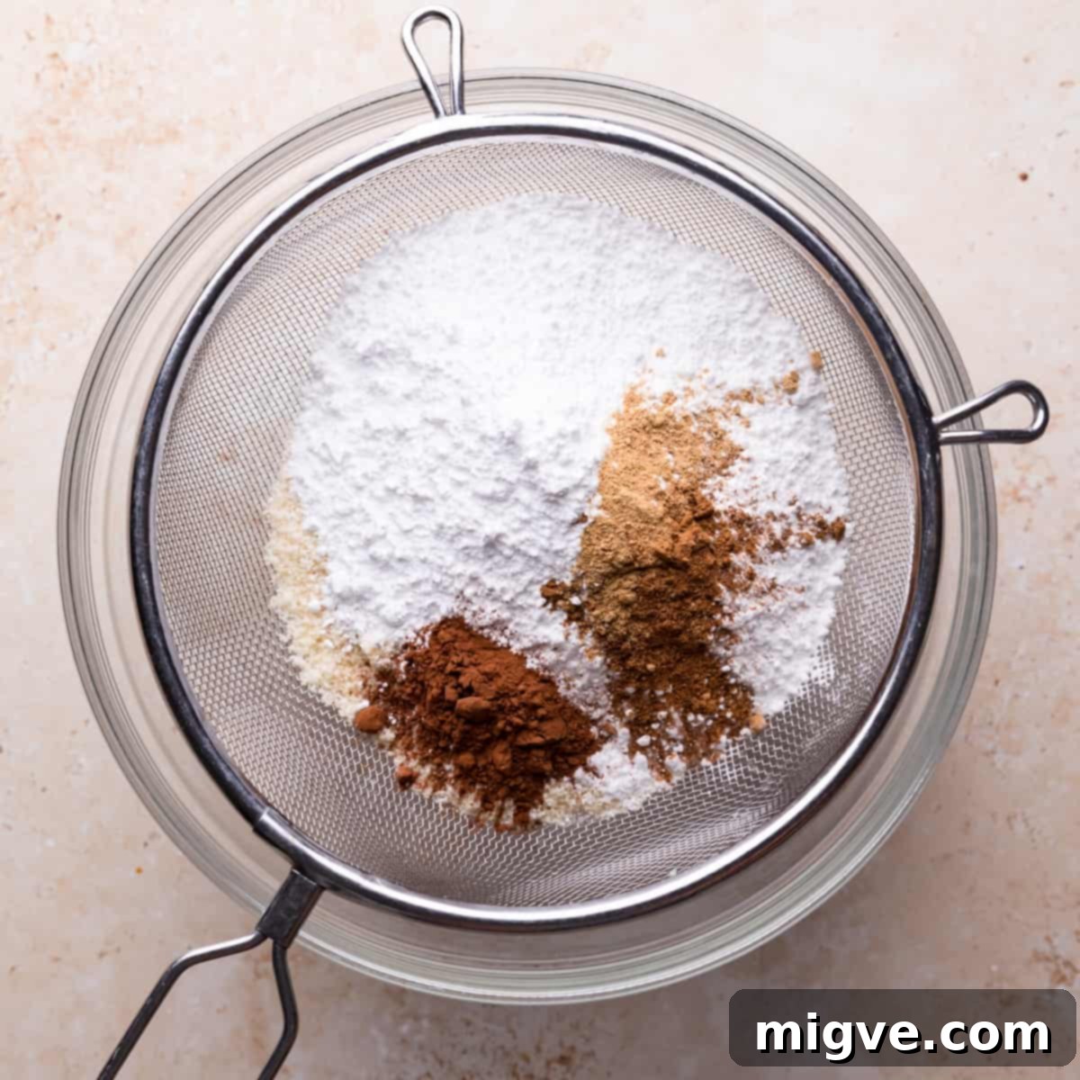A fine metal sieve filled with almond flour, powdered sugar, gingerbread spices, and cocoa powder positioned over a mixing bowl.