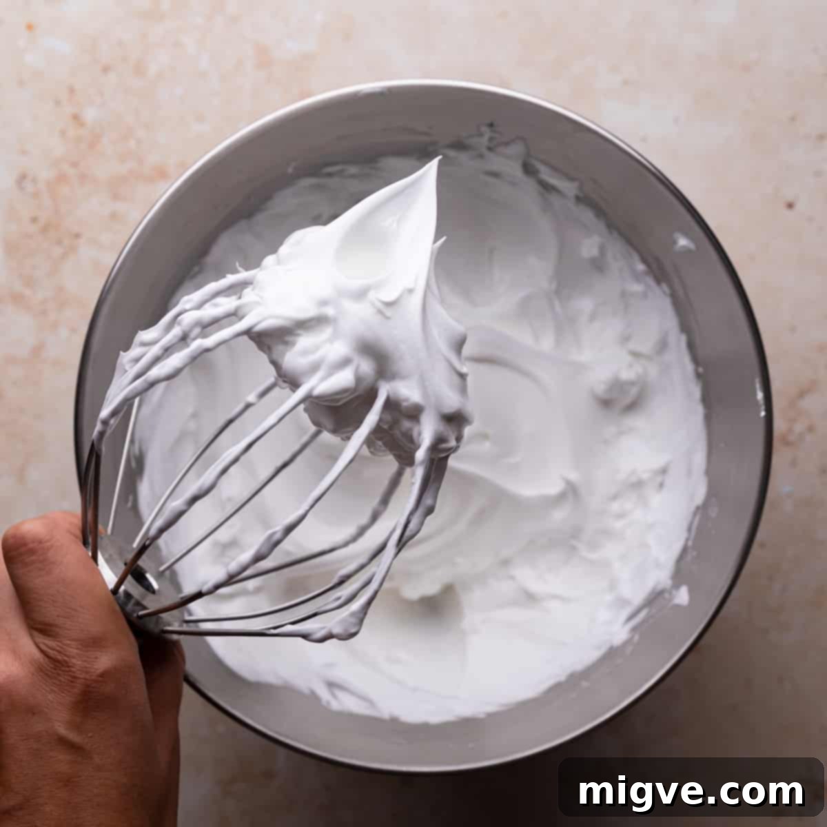 A balloon whisk held above a metal bowl, displaying stiff, glossy meringue peaks.