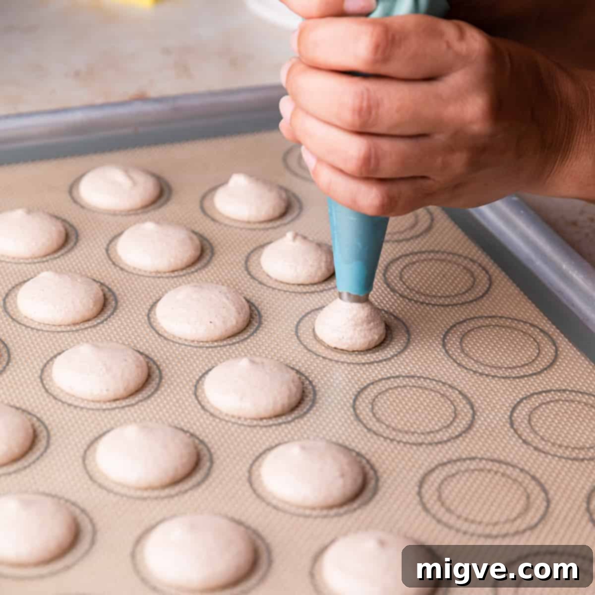 Macaron batter being piped into small, uniform circles onto a silicone baking mat with printed guides.