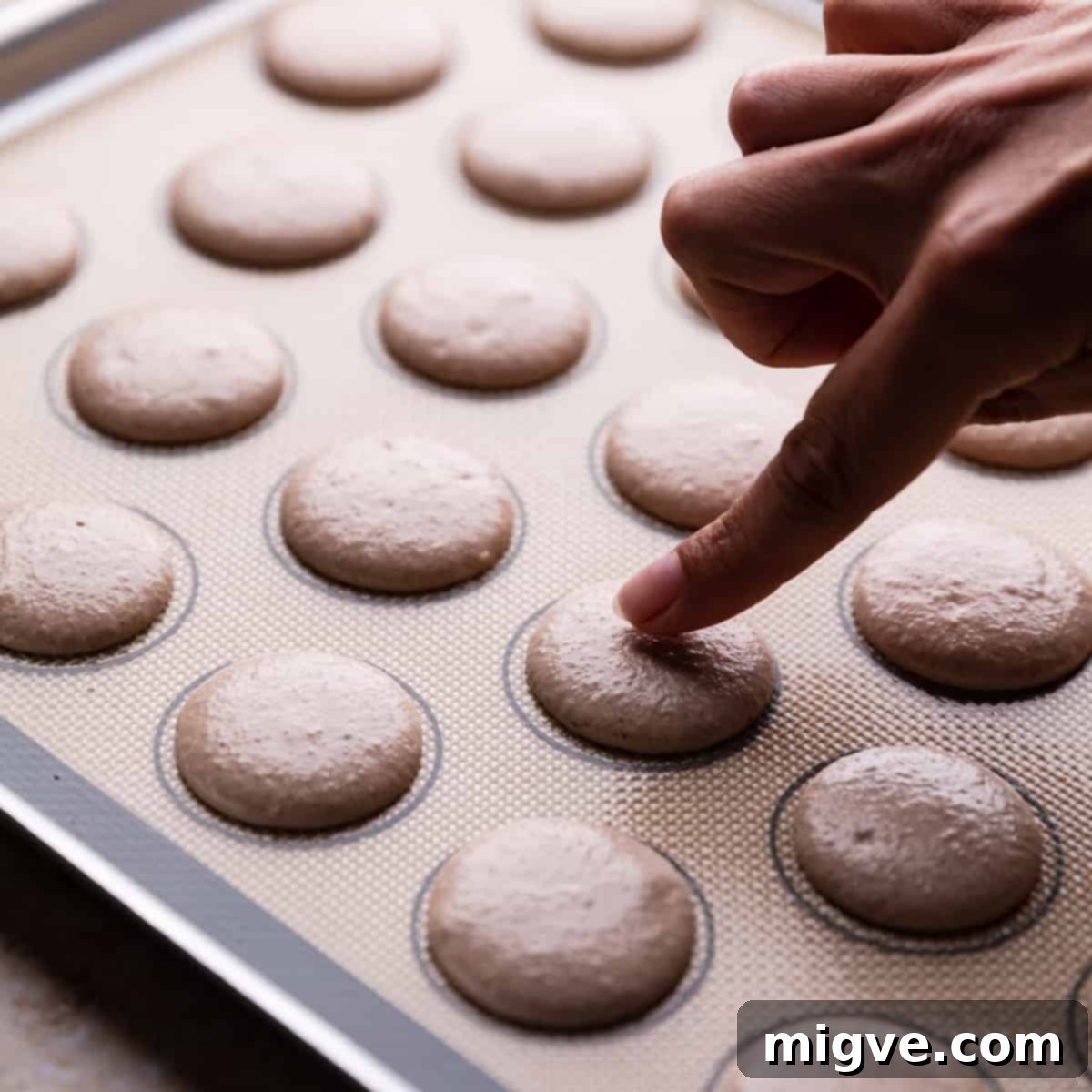 An index finger gently touching a piped gingerbread macaron on a silicone baking mat, checking for 'skin' formation.