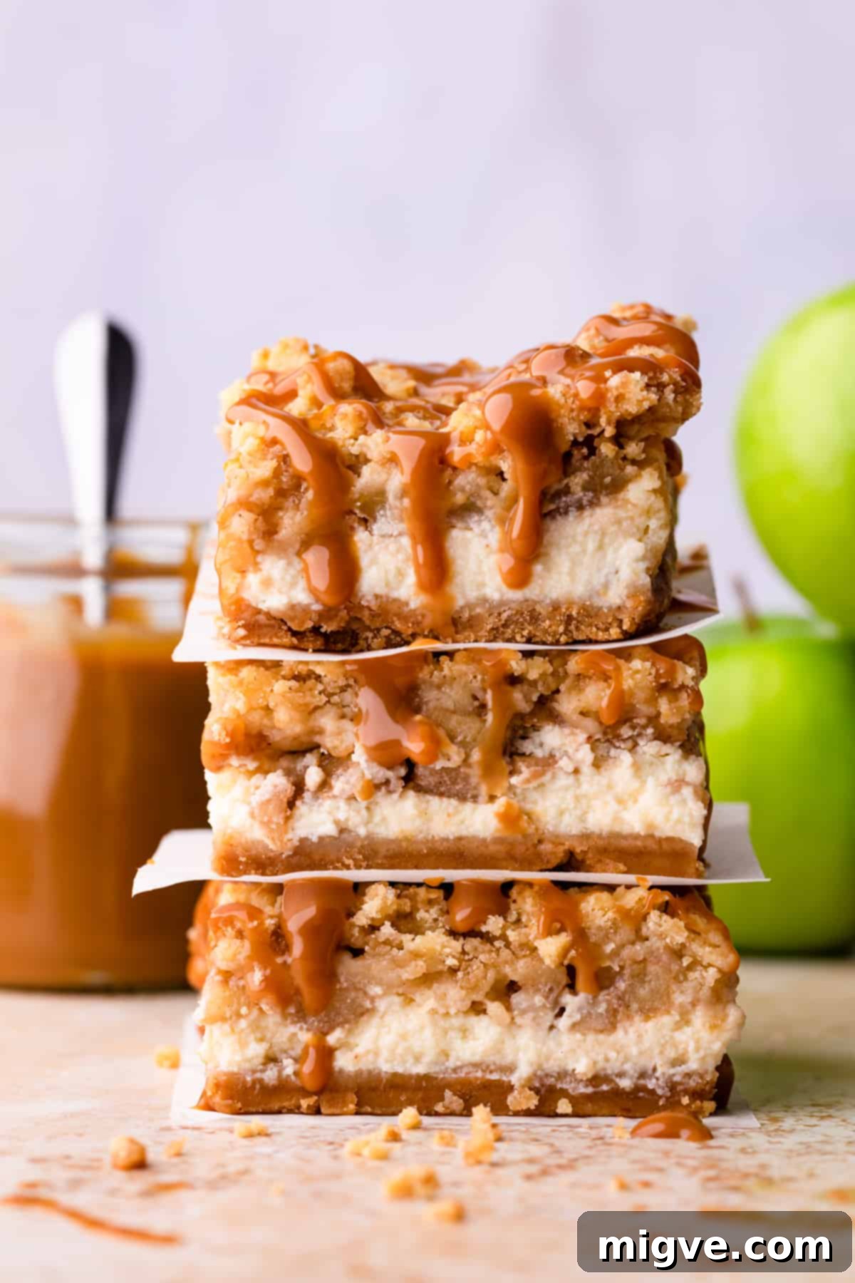 A close-up stack of three caramel apple cheesecake bars, artfully drizzled with golden caramel sauce, highlighting their multi-layered indulgence and making them appear incredibly appetizing.