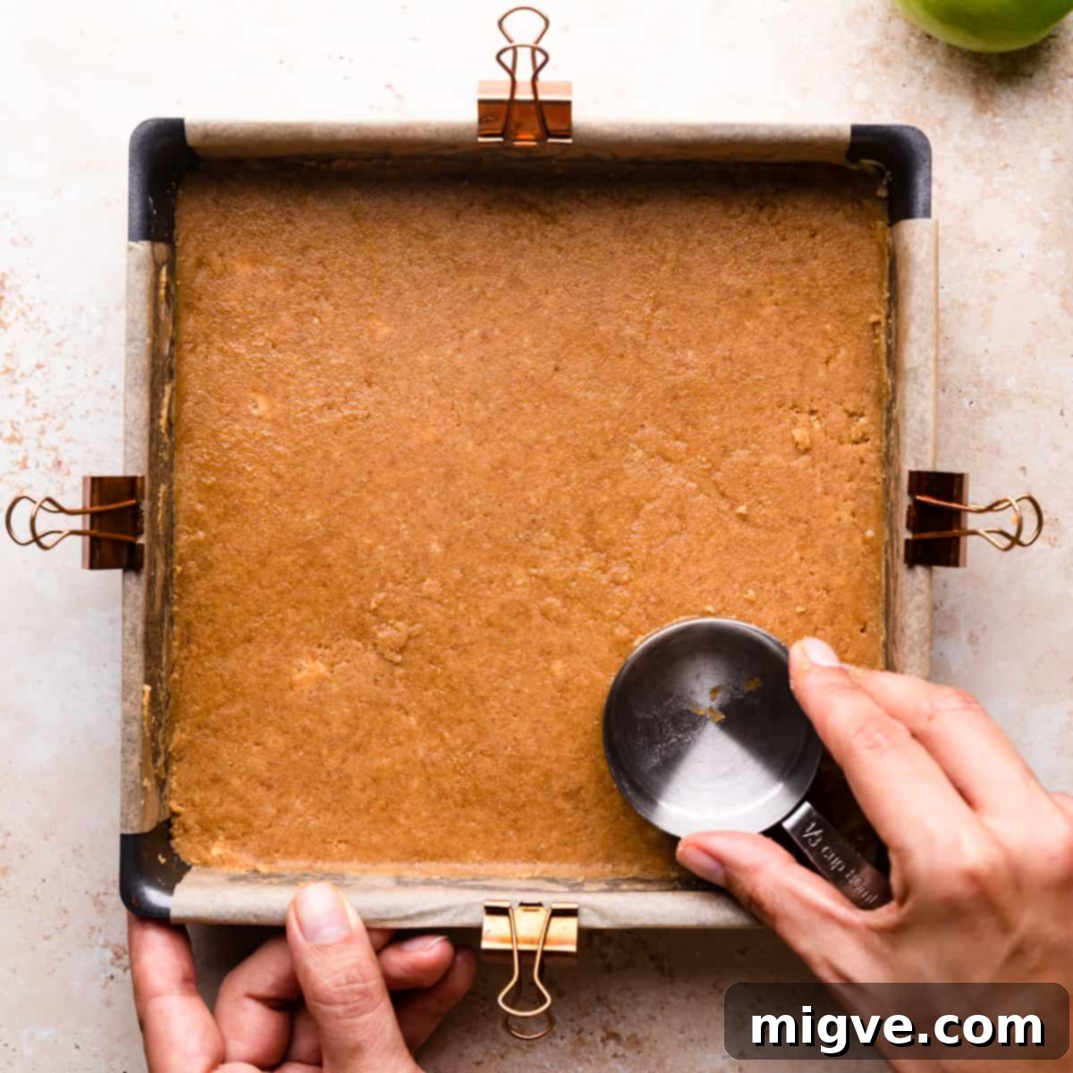 Close-up shot of a hand firmly pressing biscuit crumbs mixed with melted butter into the base of a lined baking tin, creating an even and compact crust for cheesecake bars.