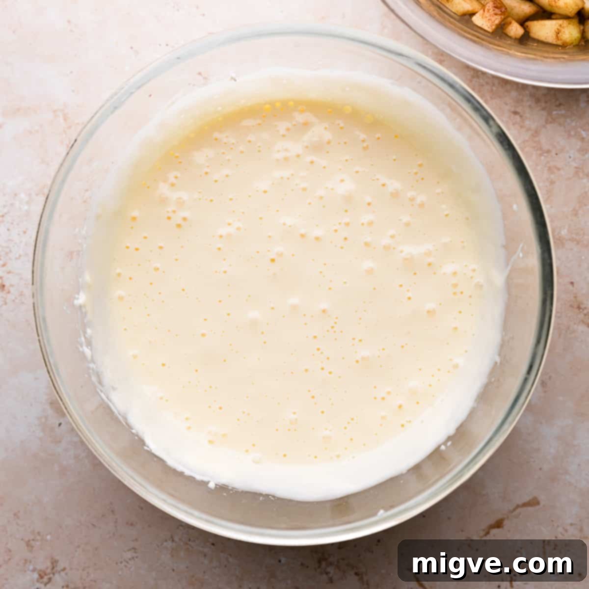 A clear glass bowl filled with smooth, pale yellow cheesecake batter, ready to be poured over the crust for caramel apple cheesecake bars.