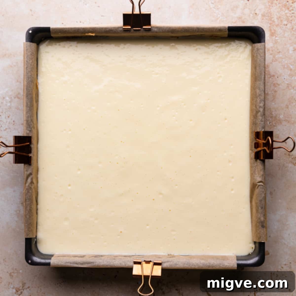 A baking pan lined with parchment paper, containing a perfectly chilled biscuit crust, topped with a smooth and creamy layer of cheesecake batter, ready for the next step.