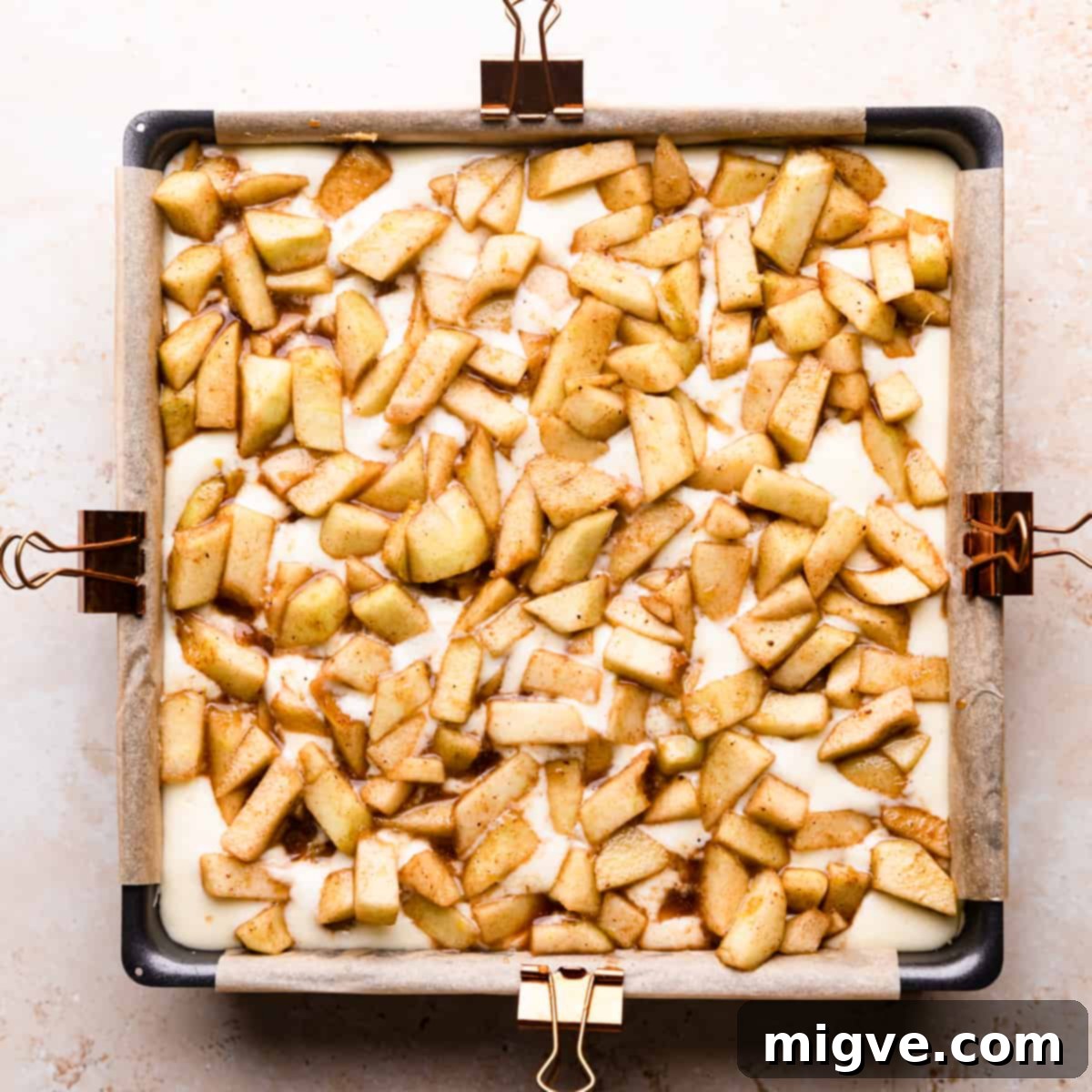 An overhead shot of a square baking pan with a creamy cheesecake layer already poured over the crust, now topped with a generous and even layer of spiced, chopped apples, ready for the crumble.