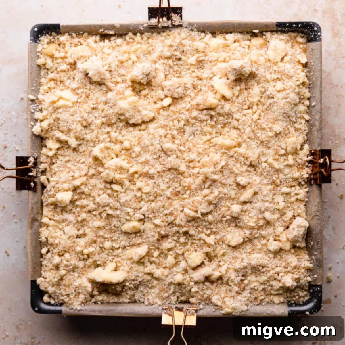 A square baking pan containing the full assembly of caramel apple cheesecake bars, with a uniform layer of golden crumble topping scattered over the spiced apples, ready to be baked to perfection.