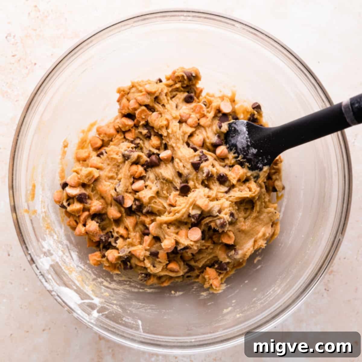 glass mixing bowl with cookie batter with butterscotch and chocolate chips inside and a rubber spatula inside of the bowl.