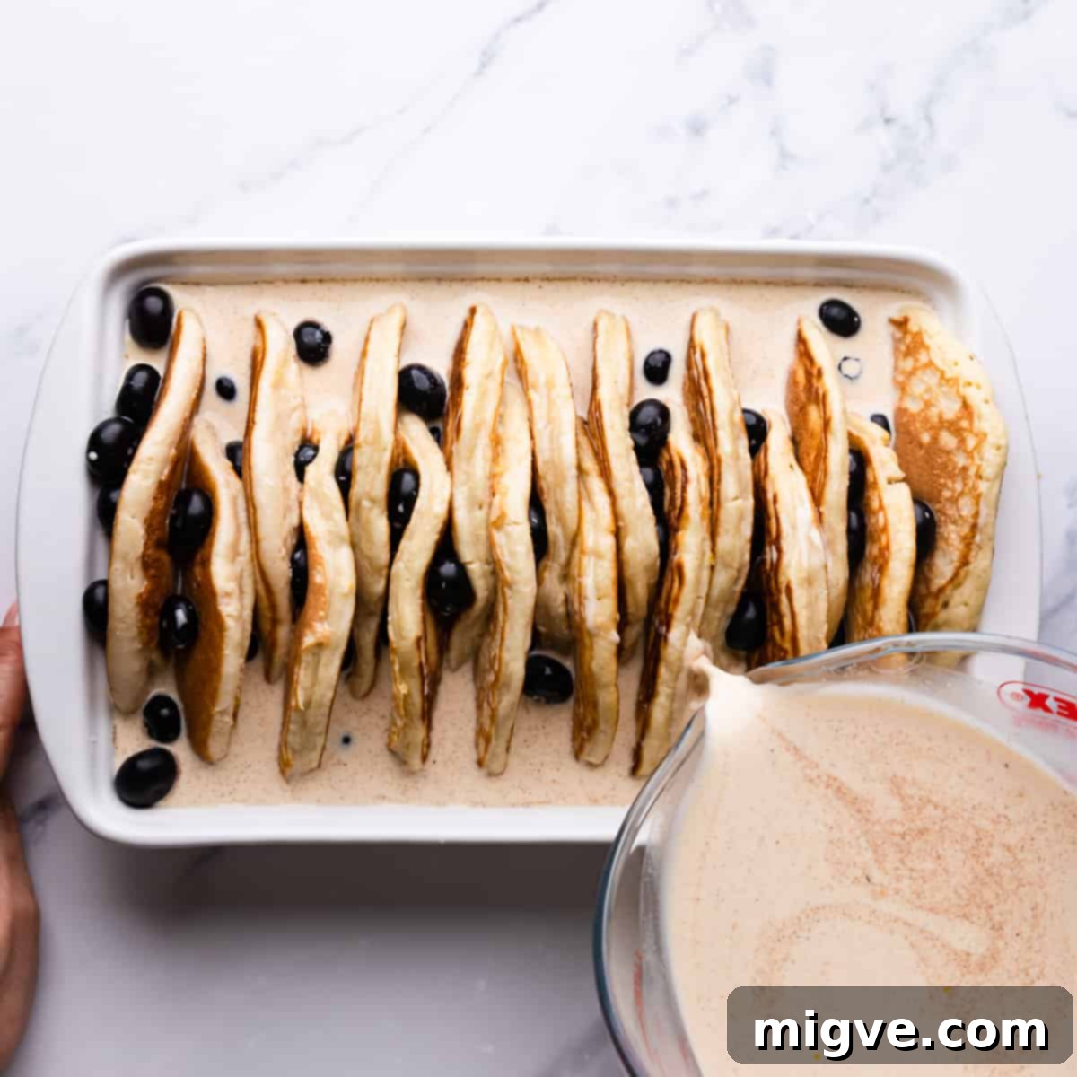 Baked Blueberry Pancake Bliss 7 Creamy custard mixture being poured over arranged pancakes and blueberries in a baking dish.