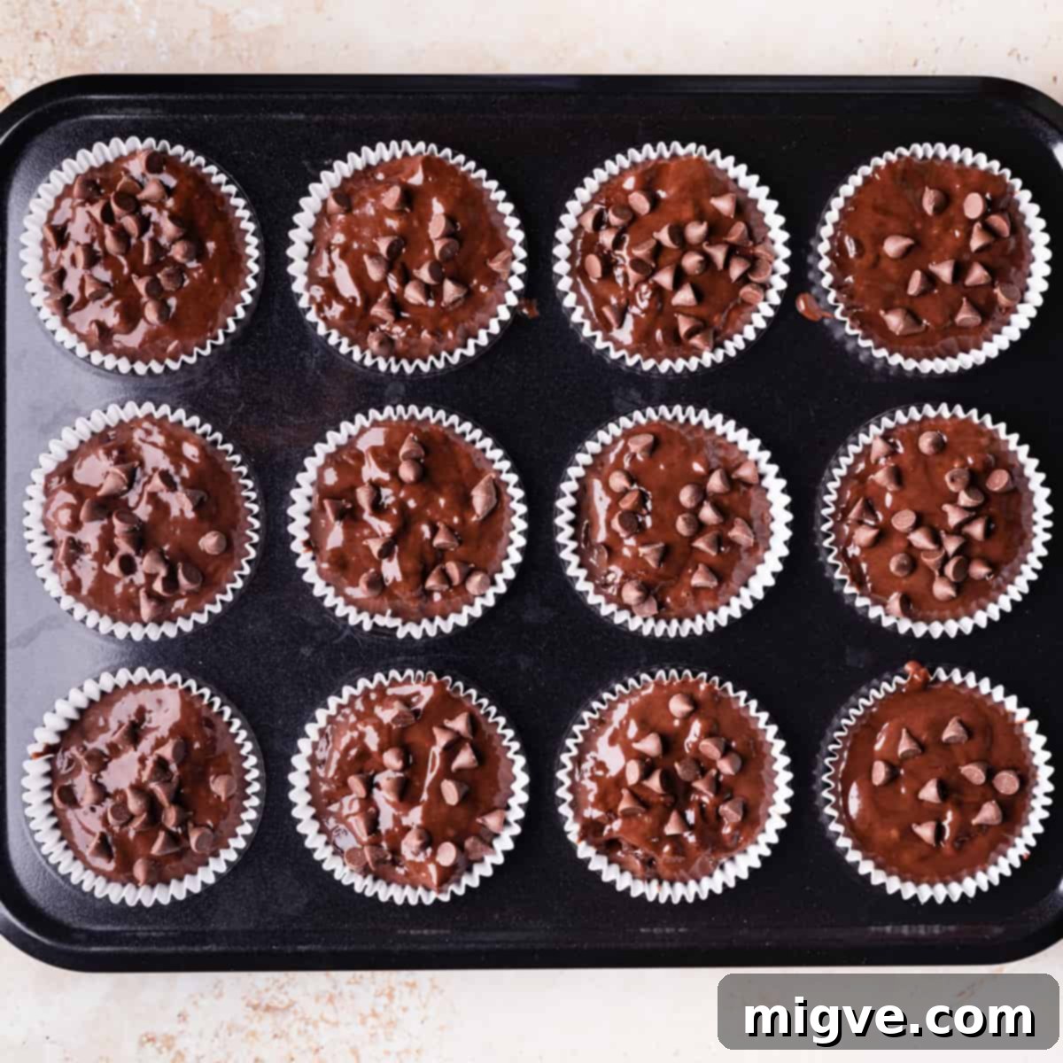A muffin pan neatly filled with chocolate muffin batter, each cup generously topped with extra chocolate chips.