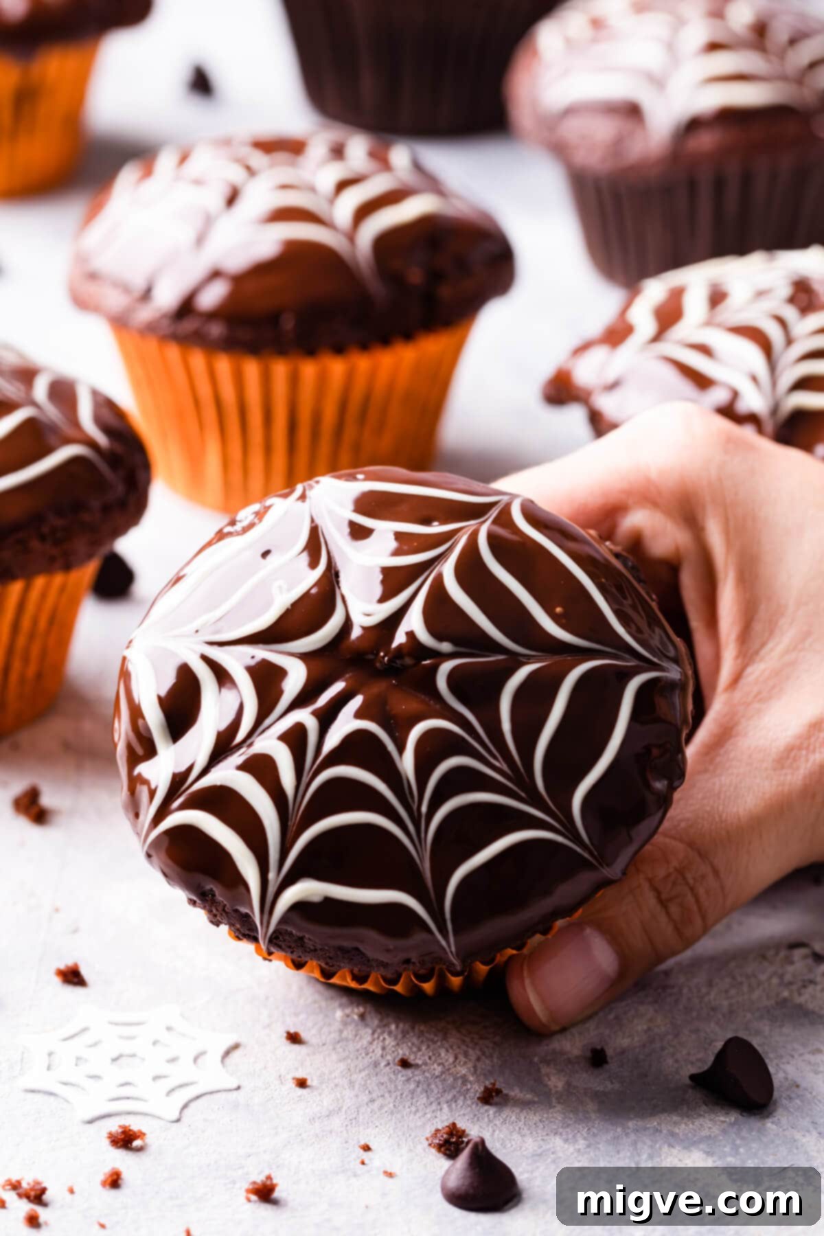 A hand holding a single chocolate fudge spiderweb muffin with more muffins blurred in the background, showcasing the moist texture and spooky decoration.