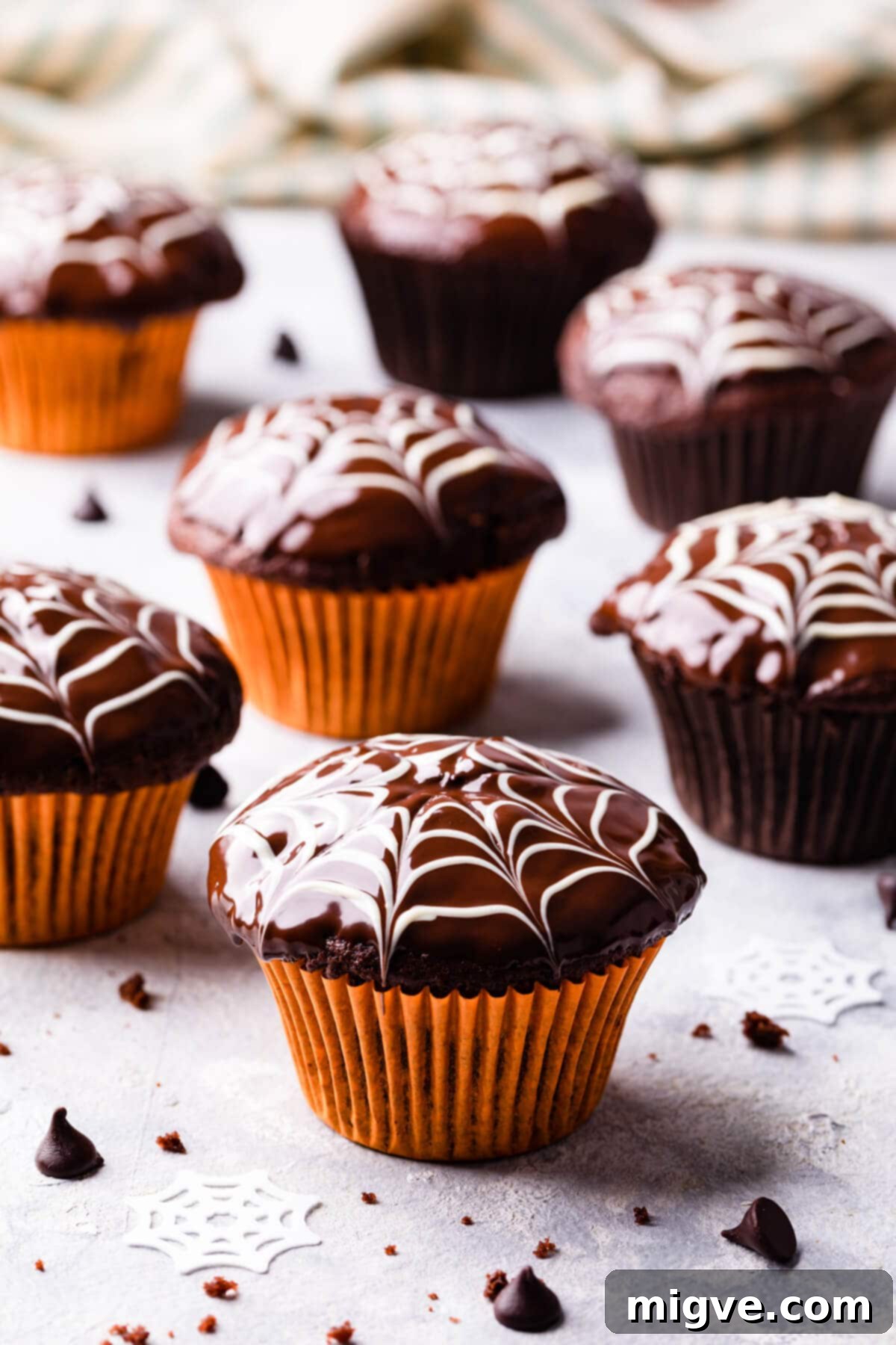 A group of beautifully decorated chocolate fudge spiderweb muffins resting in vibrant orange muffin cases on a light-colored surface.