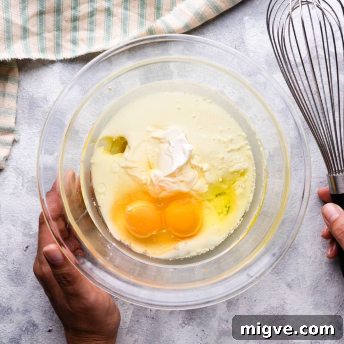 A medium glass bowl containing the whisked wet ingredients for the chocolate muffins, with a hand holding a whisk nearby.