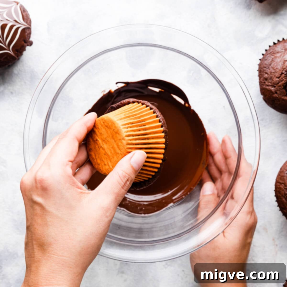 A pair of hands carefully dipping a cooled chocolate muffin into a bowl of glossy melted dark chocolate.