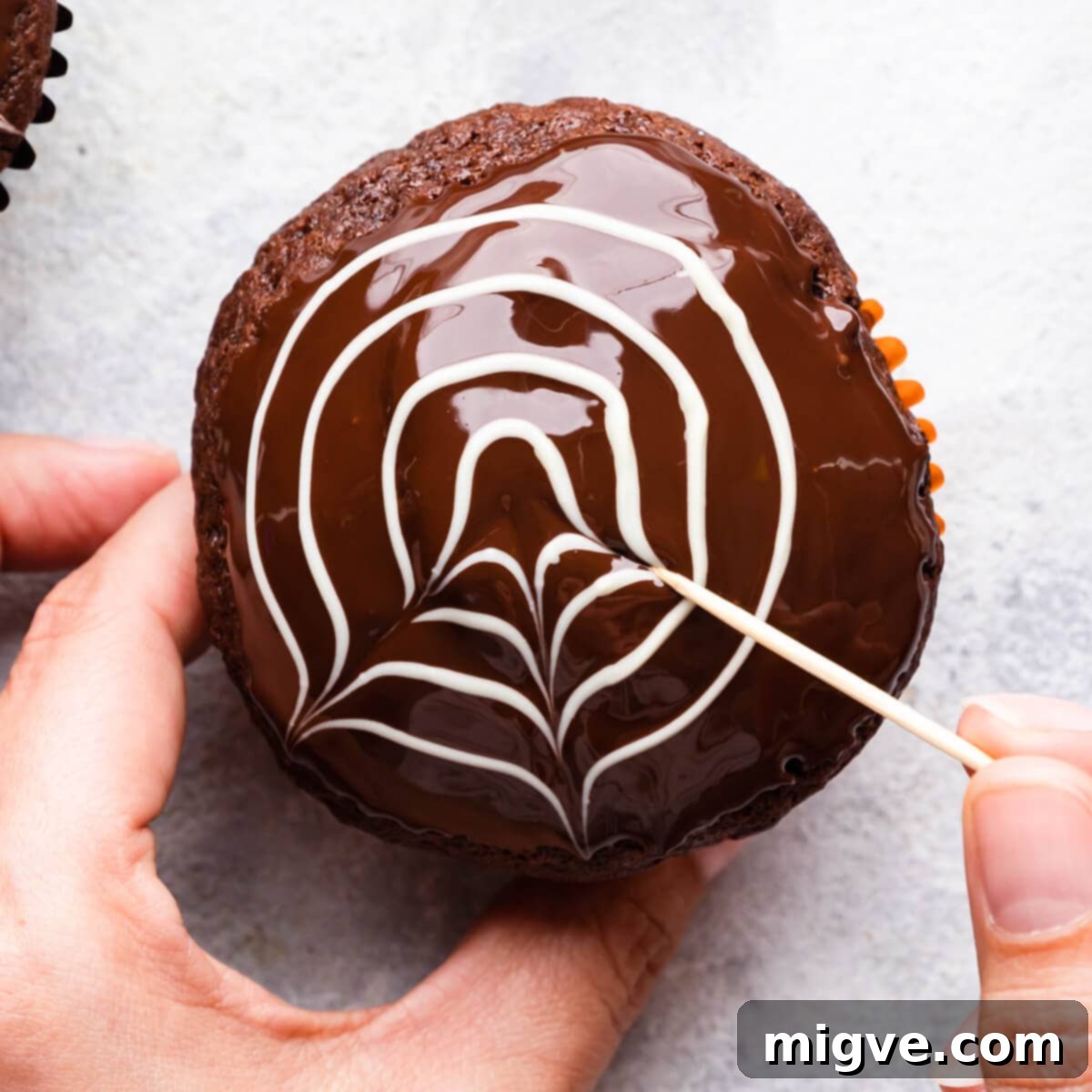 A close-up of a chocolate muffin being decorated, with a toothpick dragging through white chocolate circles to start creating the spiderweb design.