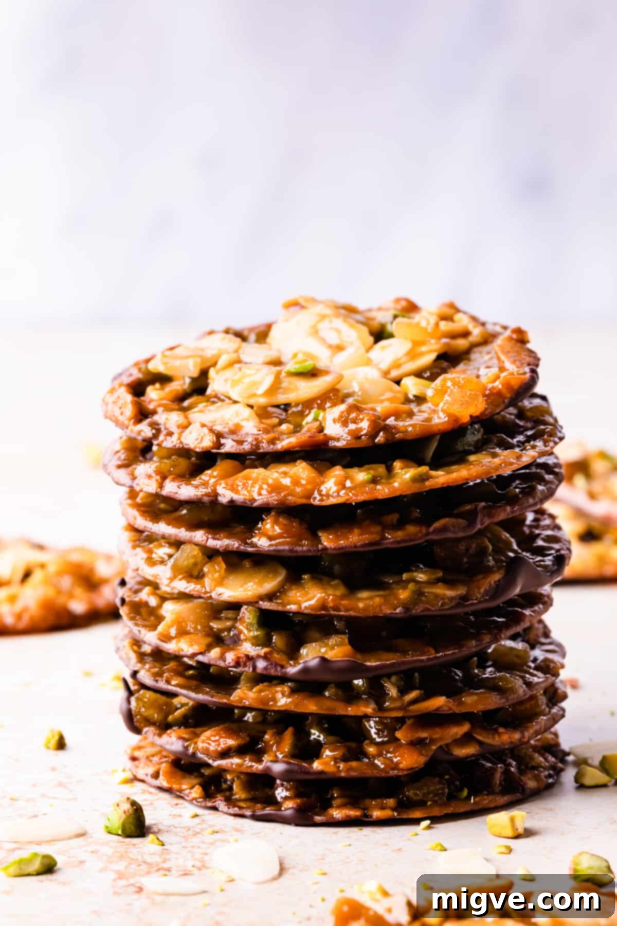 Florentine Almond Lace 12 Stack of vegan florentines with almonds and pistachios, surrounded by scattered chopped nuts, highlighting their delicious texture.
