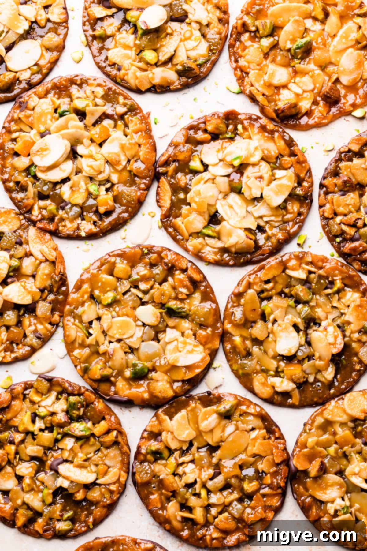 Florentine Almond Lace 18 Batch of florentine cookies with almonds on a bright surface, surrounded by scattered flaked almonds and chopped pistachio crumbs, ready for serving.