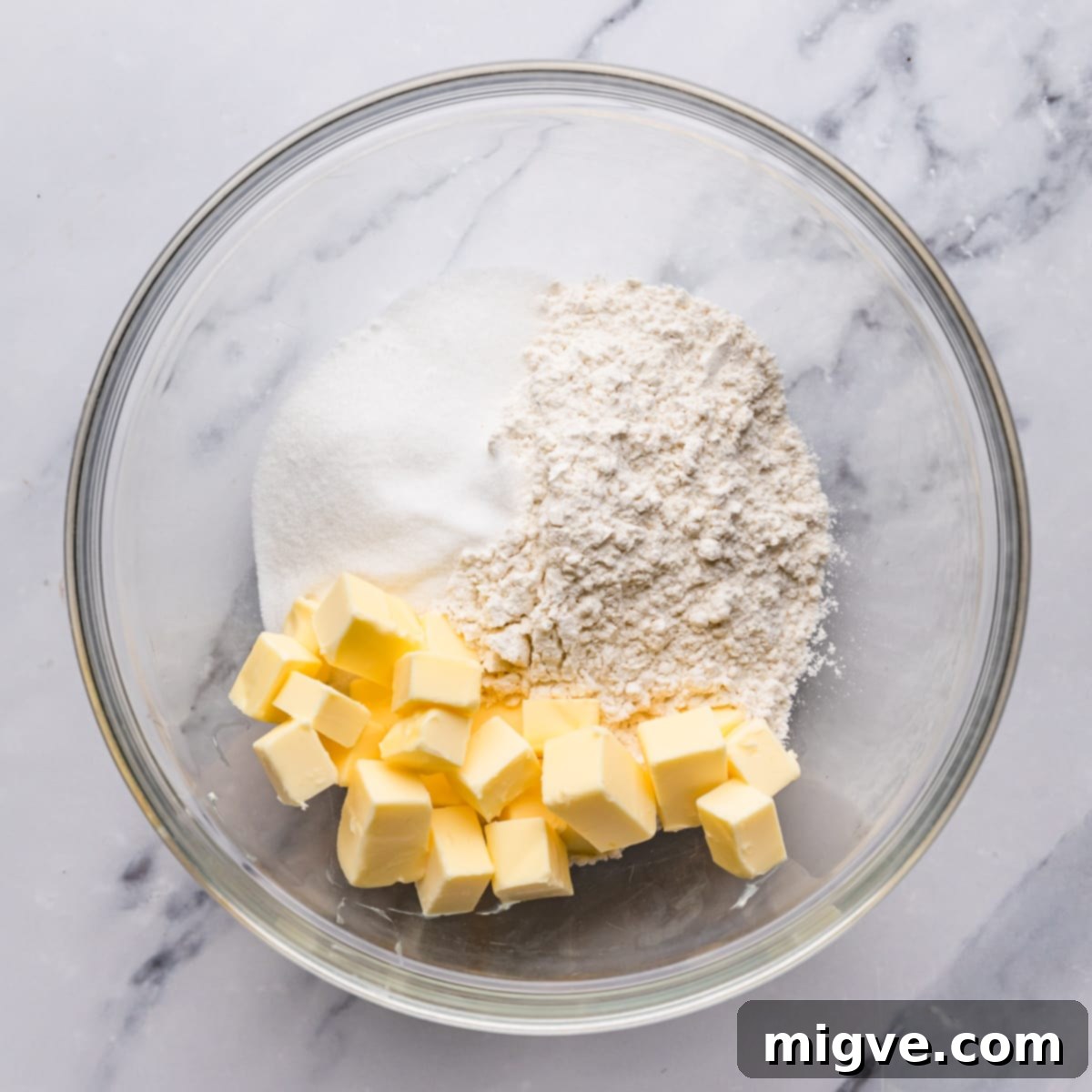 A large glass mixing bowl containing flour, butter, and sugar, ready to be combined for shortbread dough.