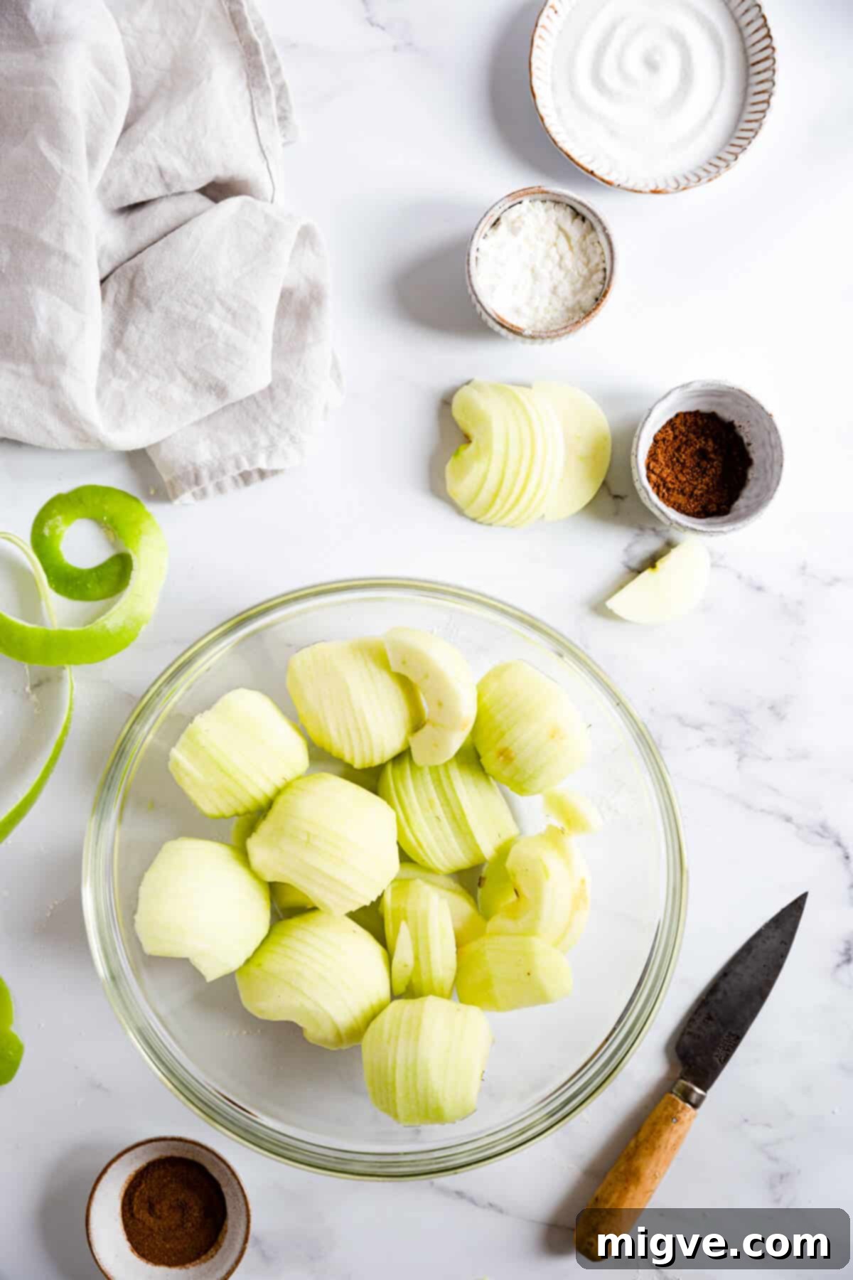 An overhead shot of a large glass bowl filled with neatly sliced, fresh apples, ready to be seasoned for the pie filling.