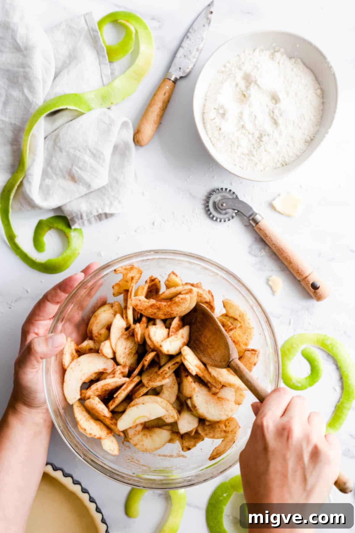 An overhead view of a person gently mixing sliced apples with a blend of spices in a large glass bowl, preparing the flavorful pie filling.